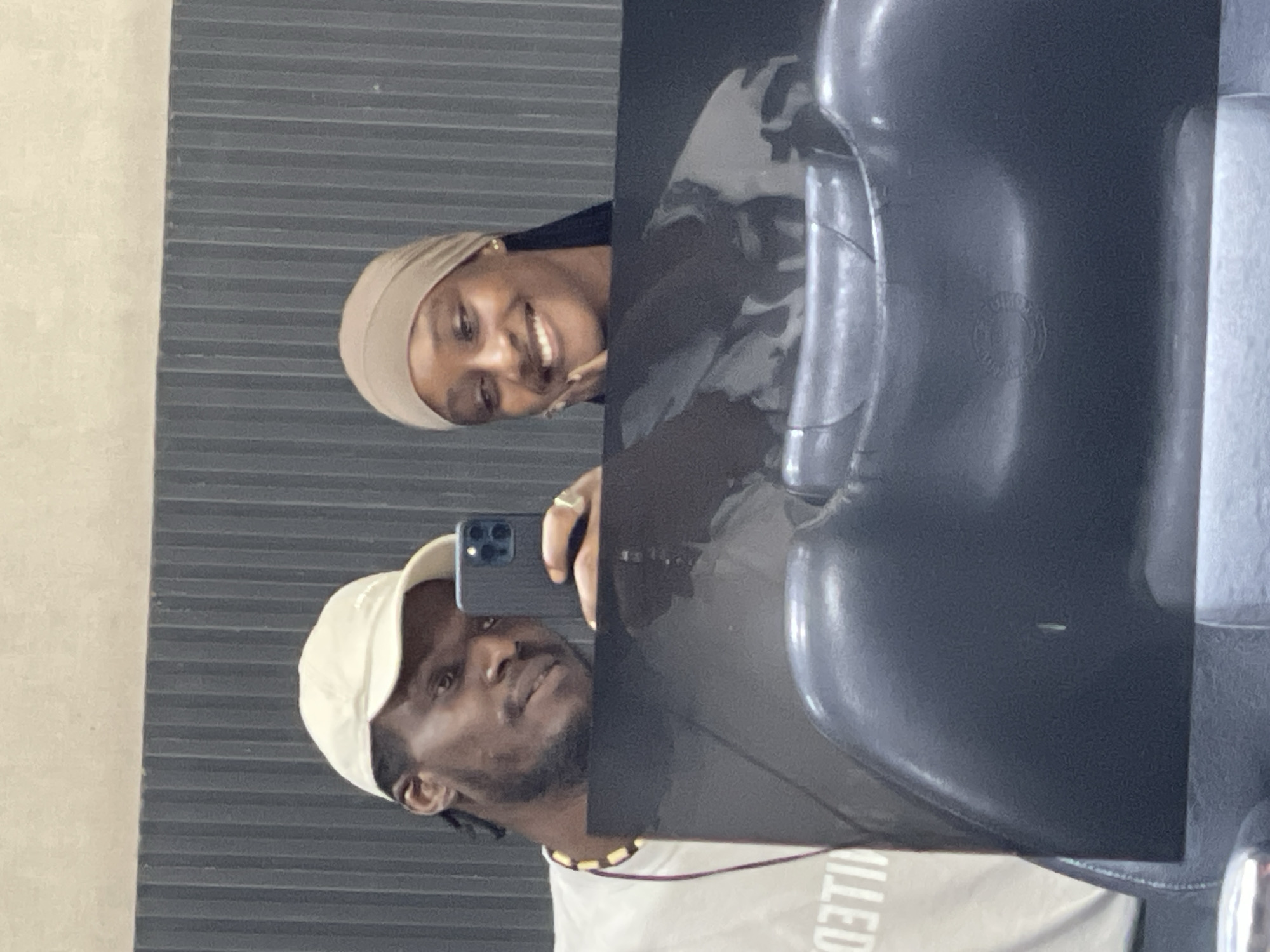 In a well-lit indoor setting, possibly a salon or studio in Guzape, Nigeria, a young African couple takes a mirror selfie. The man, wearing a light-colored baseball cap and a t-shirt with a printed design, holds a smartphone to capture the reflection. To his right, a woman in a tan headscarf smiles warmly, resting her chin on her hand. They are positioned in front of a dark, ribbed wall. In the foreground, the reflection shows the backs of two salon chairs, one dark and smooth, the other a lighter, possibly metallic shade. The overall impression is one of casual intimacy and personal documentation.