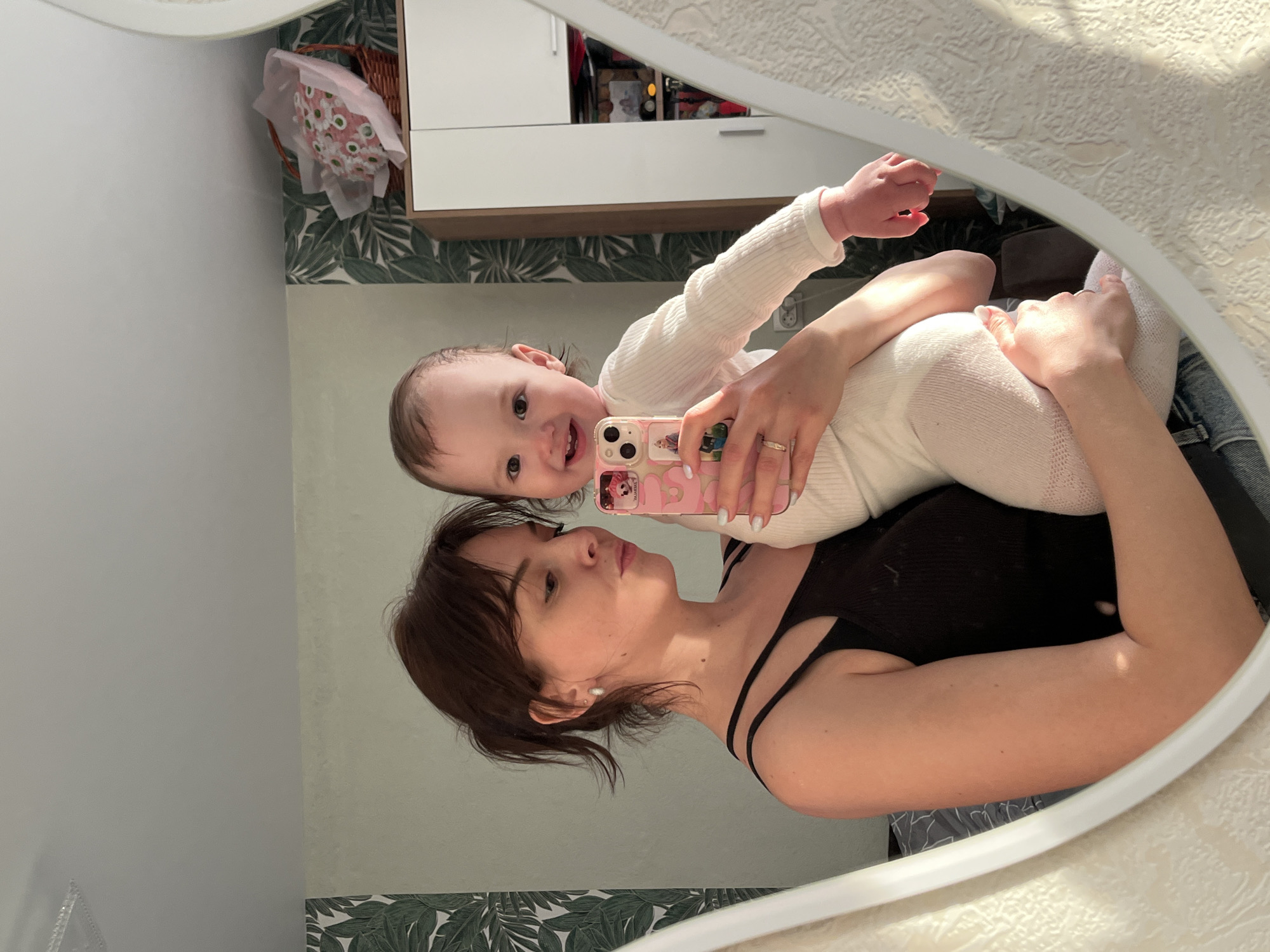 A reflection of a woman holding a baby in front of a mirror. The woman is wearing a black tank top, and the baby is wearing a white onesie and tights. The woman is holding a pink phone with a cartoon character on it. The background includes a white cabinet, a basket with flowers, and wallpaper with green leaves. The location could be Saint Petersburg, Russia based on the architecture and style of the interior.