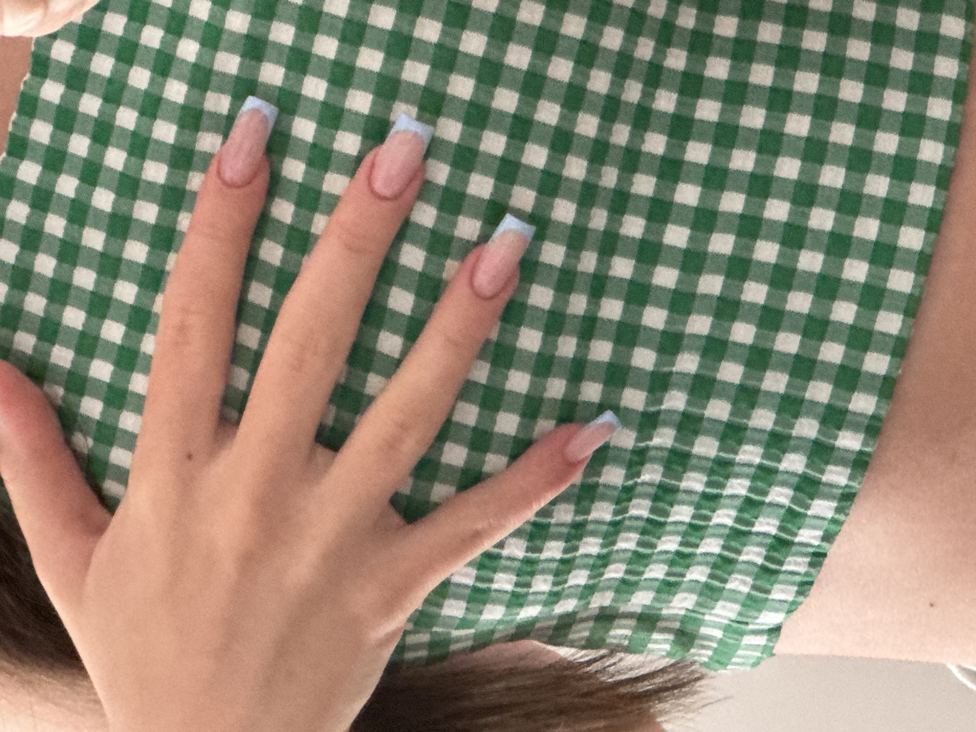 The media file shows a close-up view of a person's hand with manicured nails resting on a green and white checkered fabric. The fabric appears to be a tablecloth or a piece of clothing with a gingham pattern. The hand has long, artificial nails with a French manicure style, featuring a natural pink base and light blue tips. The skin tone of the hand is fair. The background is mostly occupied by the checkered fabric, with a hint of darker hair visible at the bottom of the frame, suggesting the person is likely sitting or leaning against something. The lighting appears to be natural, and there are no clear indications of the time of day, weather, or specific location. The overall mood conveyed is calm and perhaps a bit casual, focusing on the aesthetic of the nails and the texture of the fabric. There is no visible text in the image.