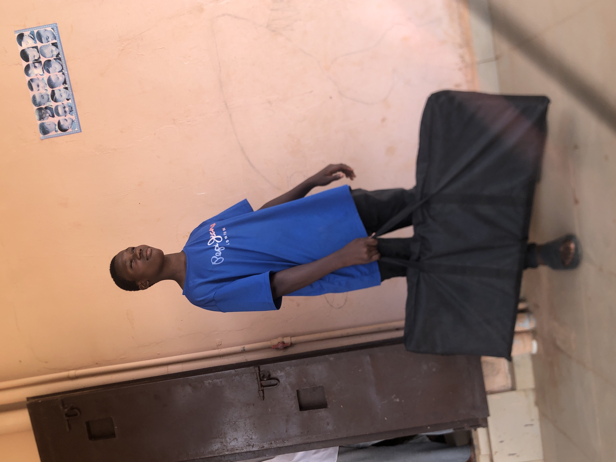 A young man wearing a blue t-shirt and black pants is standing in a hallway in Nkwelle, Nigeria, holding a large, rectangular black bag with handles. The hallway has a peach-colored wall with a row of small portrait photos attached. Below the wall is a dark metal cabinet. The floor is tiled.