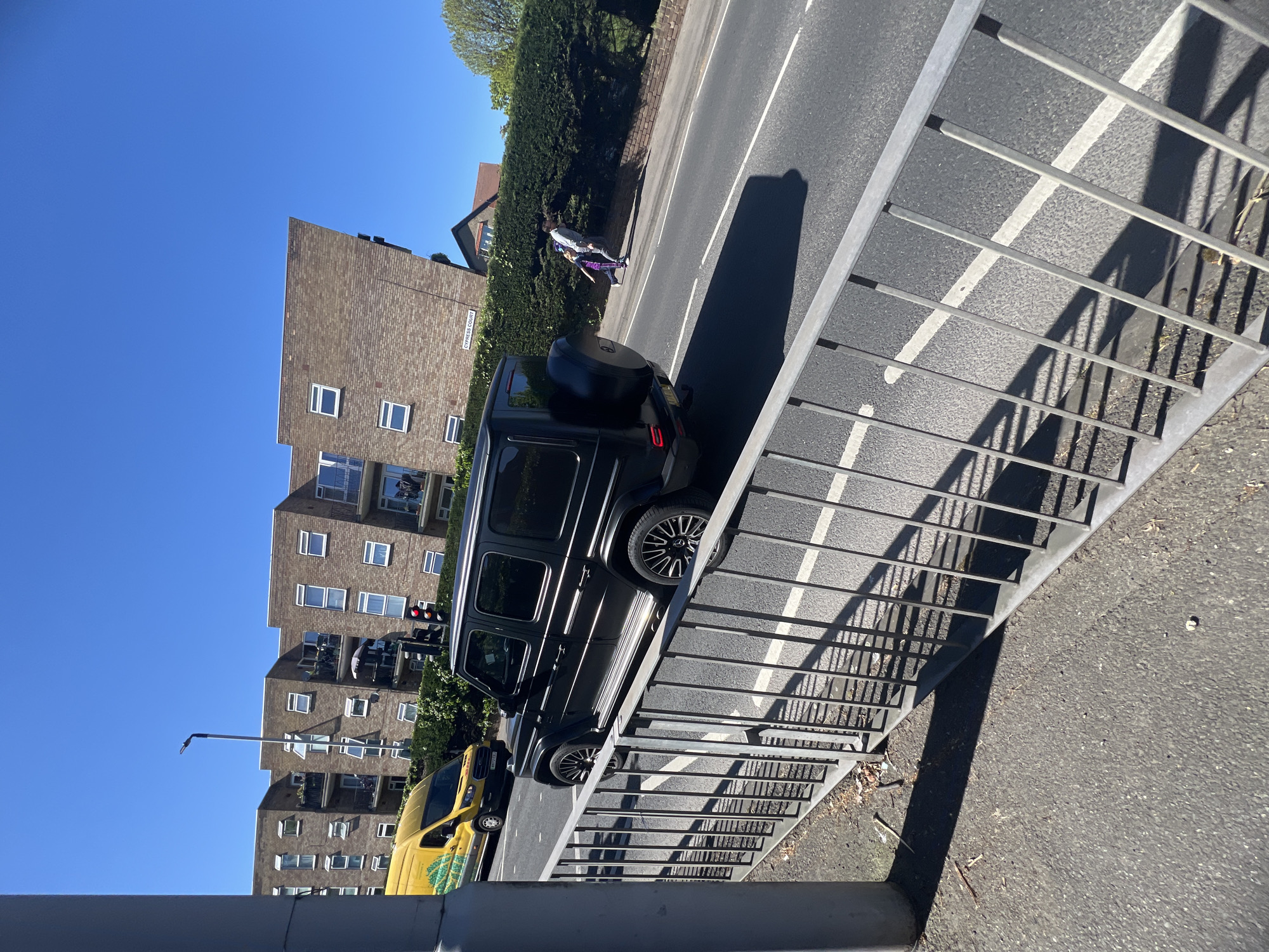 The image displays a sunny urban street scene, captured from an elevated perspective, looking down and slightly across the road. The clear blue sky dominates the upper portion of the frame.
In the foreground, a gray metal railing runs diagonally from the bottom right towards the middle left, casting long, distinct shadows onto the asphalt road and the adjacent ground. The road features clear white painted lines.
A black Mercedes-Benz G-Wagen SUV is prominently visible on the road, with its rear facing the viewer. It features a spare tire mounted on the back door and intricate silver and black wheel rims. Further down the street, a yellow Ford Transit-style van is partially visible on the left, displaying a green graphic design on its side panel. A red traffic light can be seen near the van.
Along the left side of the road, a long, well-maintained green hedge separates the street from multi-story residential buildings. A white sign reading "CYPRESS COURT" is affixed to one of the brown brick apartment blocks. Two people, an adult and a child, are walking on the sidewalk next to the hedge, further away from the black SUV. The adult wears light clothing, while the child is dressed in purple and white.
The brick buildings are modern apartment complexes with numerous windows and balconies. A street lamp post is also visible. The scene is brightly lit by the sun, casting strong shadows, suggesting it is either morning or late afternoon on a clear, cloudless day.