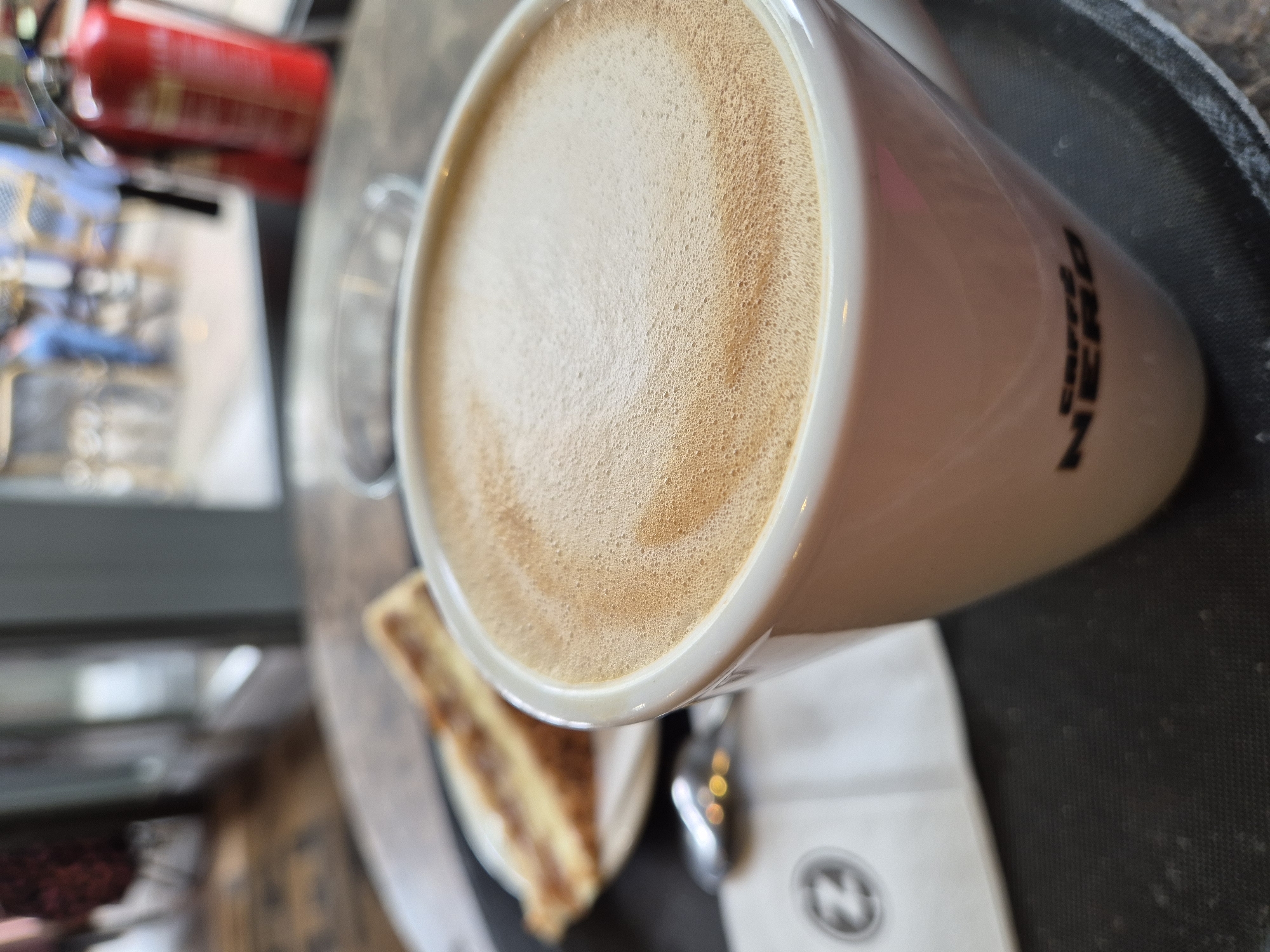 A close-up, overhead shot captures a moment of indulgence at a Caffè Nero coffee shop, likely in West Suffolk, United Kingdom. The central focus is a white ceramic mug, angled and partially rotated, filled with a creamy, frothy coffee beverage, most likely a latte or cappuccino. The surface of the coffee displays a rich, textured foam with varied shades of light brown.
The distinctive "CAFFÈ NERO" logo is clearly visible in black text on the side of the mug. Beside the mug, on a dark, textured tray, rests a small slice of a layered baked good, possibly a cake or tart, showing alternating light and dark brown layers. A white paper napkin, featuring the Caffè Nero 'N' logo within a circle, is positioned underneath the handle area of the mug, partially obscuring a metallic spoon reflecting ambient light.
The background elements are intentionally blurred, suggesting a shallow depth of field. Glimpses of a red object, possibly a fire extinguisher, and other indistinct cafe furnishings can be seen, contributing to the indoor coffee shop setting. The image evokes a sense of a relaxing break, focusing on the sensory appeal of the coffee and accompanying treat. The time of day is not discernible.