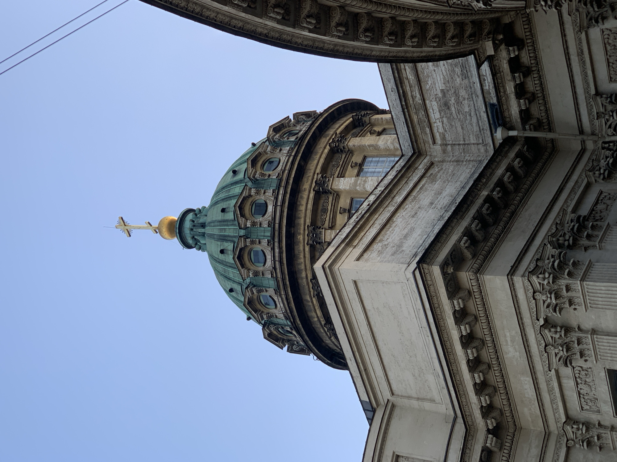 This image shows the dome of the Kazan Cathedral in Saint Petersburg, Russia, from a low-angle perspective. The dome is green, topped with a golden spire and cross. The architectural details of the cathedral are visible, including windows and ornate decorations. The sky is blue. The image is oriented vertically. Parts of other architectural structures frame the view of the dome.