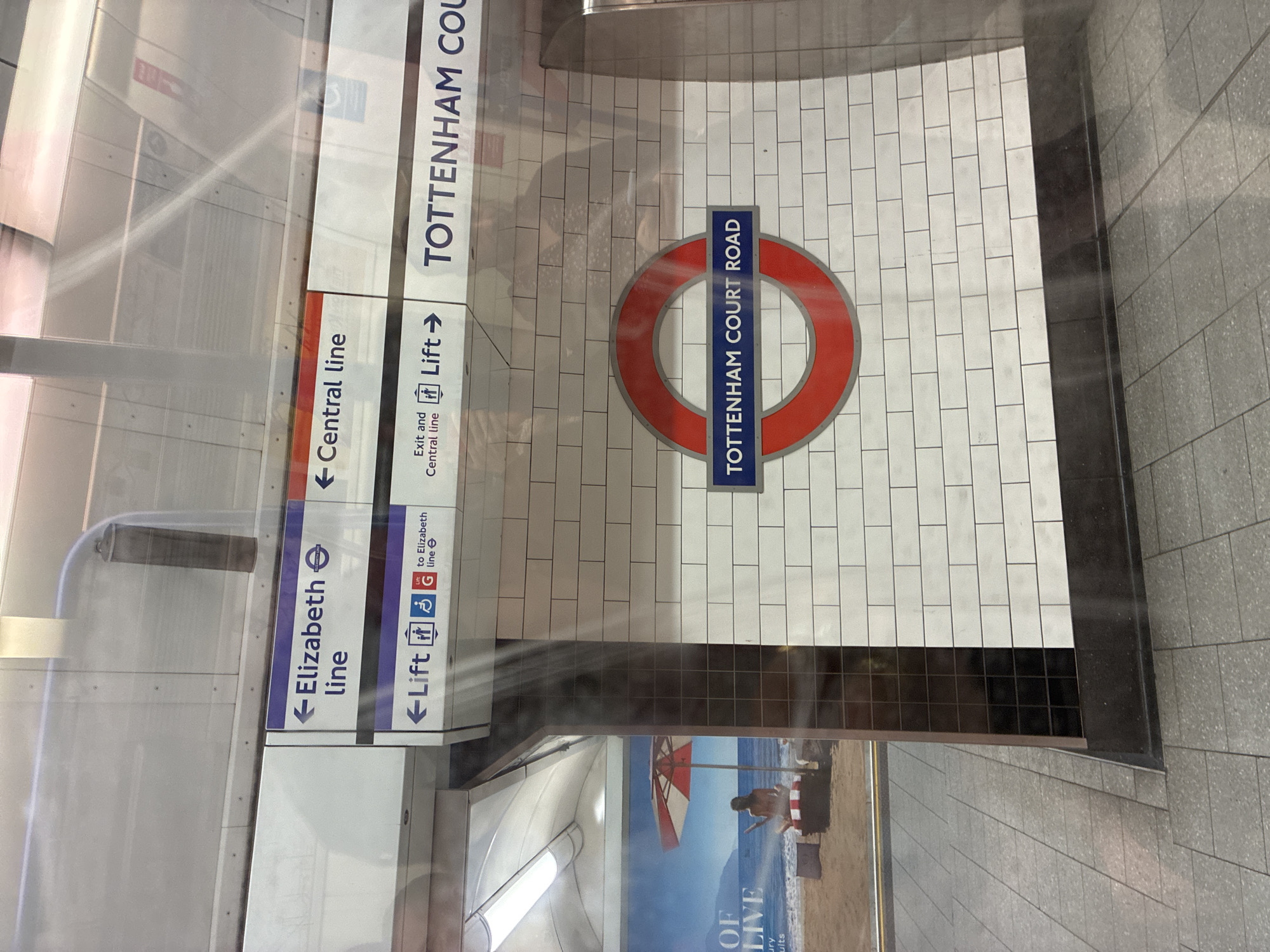 The image shows signage inside the Tottenham Court Road underground station in London, England. The signs indicate directions to the Elizabeth Line, Central Line, and lifts. A section of the wall is tiled in white, with the iconic London Underground roundel featuring a red circle with "TOTTENHAM COURT ROAD" in blue. An advertisement for a beach vacation is partially visible below the signage. The image appears to have been taken through a window or a reflective surface.