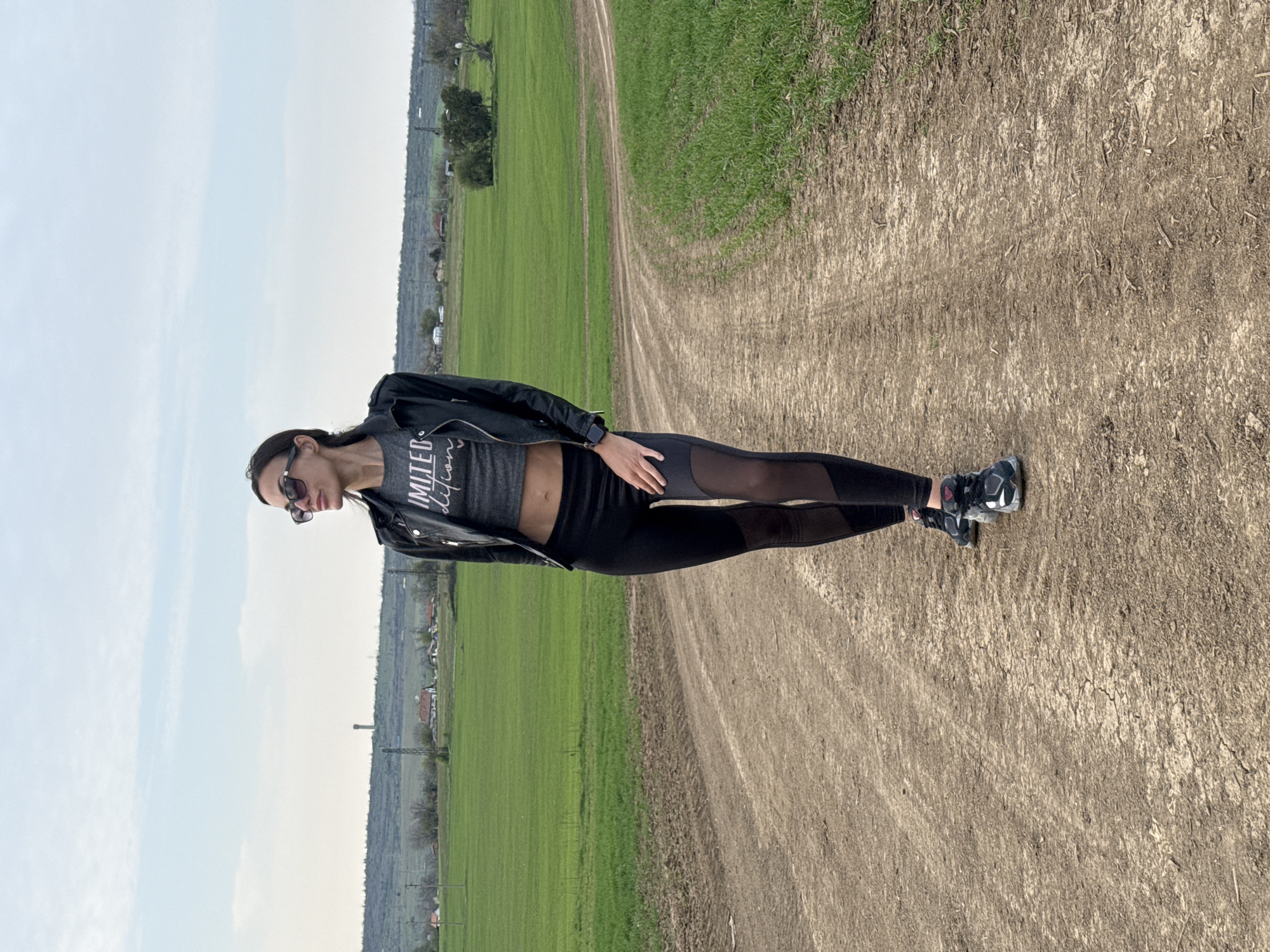 A woman stands on a dirt road in a rural area, likely in the vicinity of Pleven, Bulgaria, given the terrain and distant buildings visible in the background. She is wearing sunglasses, a gray cropped t-shirt with white text, a black leather jacket, and black leggings with sheer panels. She is also wearing athletic shoes. The dirt road has visible tire tracks and runs between fields of green grass. The sky is overcast with pale blue hues.