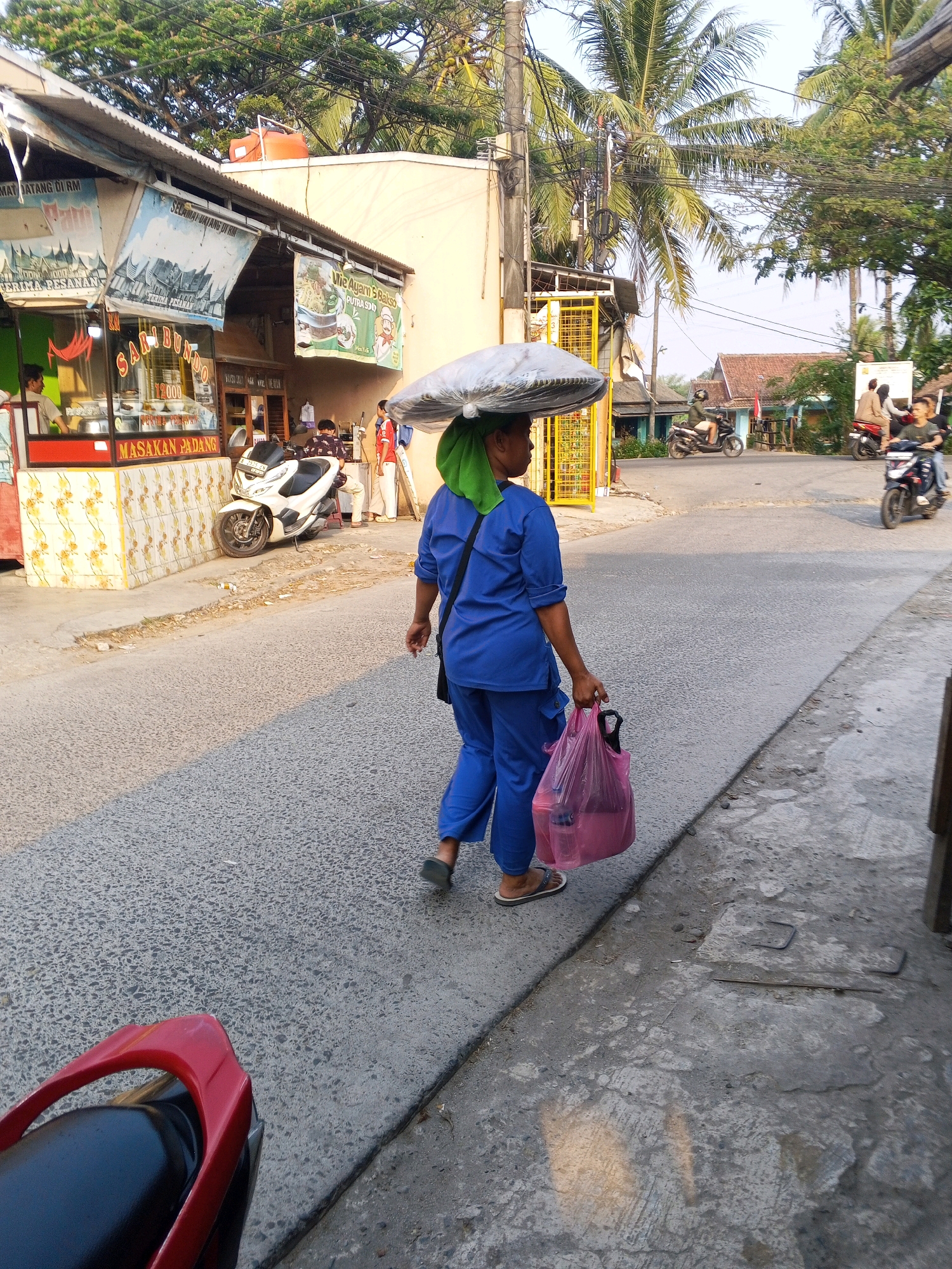A person in a blue uniform walks away from the camera on a street in Ciruas, Indonesia, carrying a pink plastic bag in their right hand and a large, flat object wrapped in gray plastic on their head. The person is wearing flip-flops. To the left of the person is a building with a sign that says "MASAKAN PADANG". A white scooter is parked in front of the building. In the background, palm trees and other vegetation are visible, as are houses and other buildings. The sky is a pale blue.