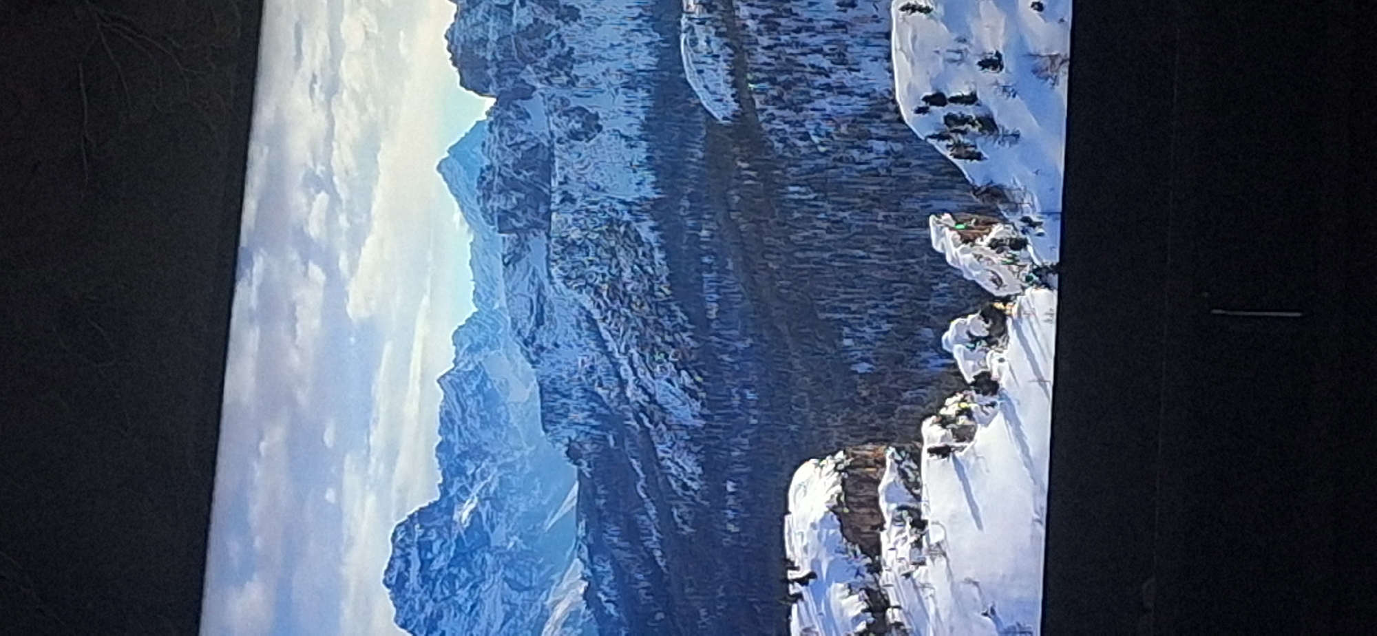 The image displays a sweeping, majestic mountain landscape covered in snow. The foreground features rugged, snow-capped rocky outcroppings, casting long shadows across the terrain. Beyond these, a dense forest of evergreen trees blankets the slopes, also dusted with snow. In the distance, a range of imposing, snow-covered mountains rises against a sky filled with soft, white clouds. The overall scene evokes a sense of vastness and natural beauty in a winter environment. There is no indication of the city of Bila Tserkva, Ukraine, in this image.