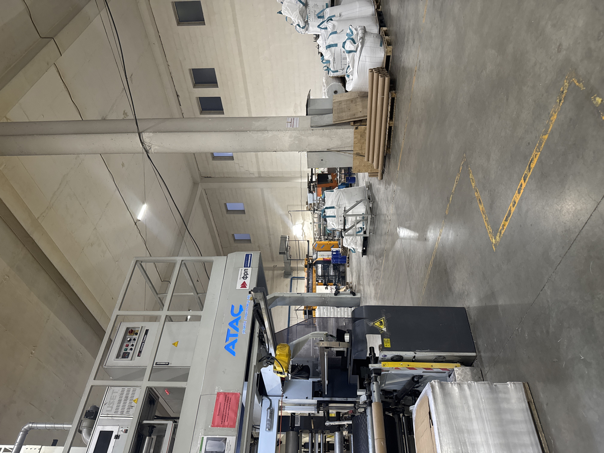 The image depicts the interior of an industrial warehouse or factory in Perm, Russia. In the foreground, a large, complex piece of machinery, likely a plastic film extrusion or printing machine, is prominent. It is gray and white with blue lettering that reads "ATAC plastic machinery co." and a logo with Russian text. To the left of this machine, a stack of wrapped materials on a pallet is visible.
The environment is a large, open space with a concrete floor marked with yellow lines, possibly indicating pathways or designated areas. In the mid-ground and background, other industrial equipment and machinery are present, including what appear to be more printing or processing machines in yellow and orange.
Towards the right side of the image, several large white sacks, likely containing raw materials or finished products, are stacked on pallets. These sacks have blue trim and some have Russian text printed on them, including what appears to be "АЛЬФАПОЛ" (Alfapol). Rolls of cardboard tubes are also stacked on a pallet nearby.
The ceiling is high and made of concrete. A large concrete pillar supports the ceiling. Natural light enters from windows high on the back wall, and artificial lighting from fluorescent fixtures on the ceiling is also present. No people are visible in the scene. The overall impression is one of an active, functional industrial setting.