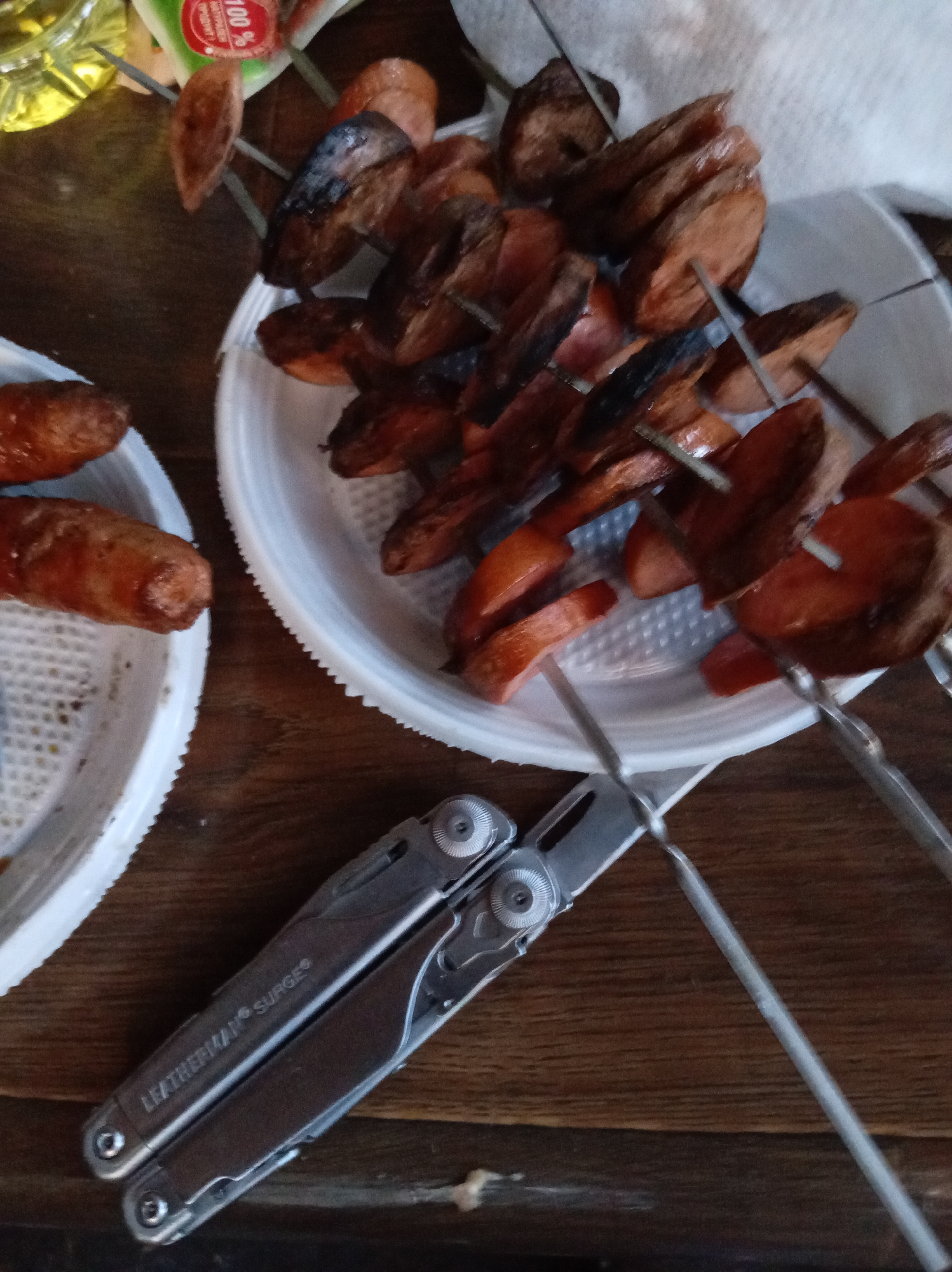 This is a close-up, high-angle shot of a wooden table, on which are placed two white paper plates filled with sliced, grilled sausages. One plate is on the left, and the other is on the right and slightly behind the first. The sausages on the right are skewered on metal rods and appear to be more thoroughly cooked, with some charring visible. A multi-tool, identified by the logo "LEATHERMAN SURGE," is positioned in the foreground, resting on the table in front of the plates. In the upper left corner, a portion of a clear bottle containing a yellow liquid and a white and green label with red lettering is visible. The environment suggests an informal meal or gathering.