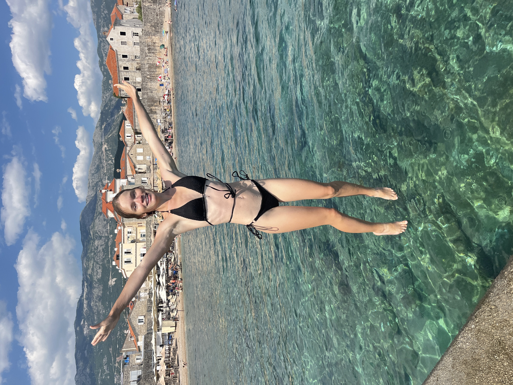 A young woman with blonde hair, wearing a black bikini, is captured mid-air with her arms outstretched, appearing to jump into the clear turquoise water. She is positioned in the foreground, with the city of Budva, Montenegro, in the background. The city features buildings with red-tiled roofs and stone walls, situated along a coastline with a beach dotted with umbrellas and people. Mountains form a backdrop to the urban landscape, and the sky is blue with scattered white clouds. The water is transparent, allowing a view of the rocky seabed. The image appears to be taken from a slightly low angle, emphasizing the woman's jump.