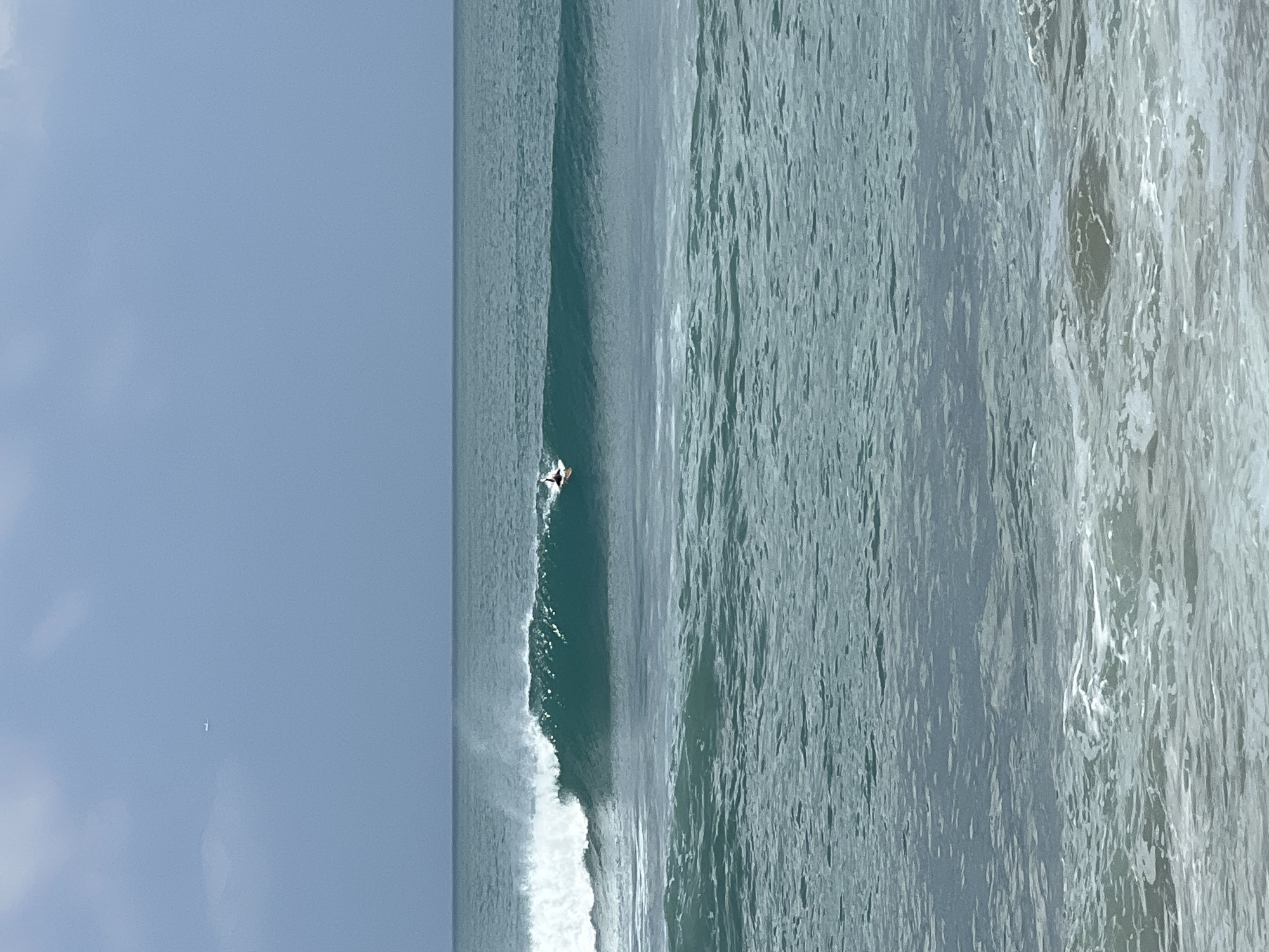 A surfer rides a wave in the ocean off Kuta, Indonesia. The surfer is positioned on the right side of the image, riding the crest of a turquoise wave. The wave is breaking to the left, with white foam trailing behind it. In the distance, the ocean extends to the horizon, meeting a hazy blue sky. A small, distant airplane is visible in the upper left quadrant of the sky. The water in the foreground is choppy, with smaller waves and whitecaps. The scene captures a moment of surfing activity in a coastal environment.