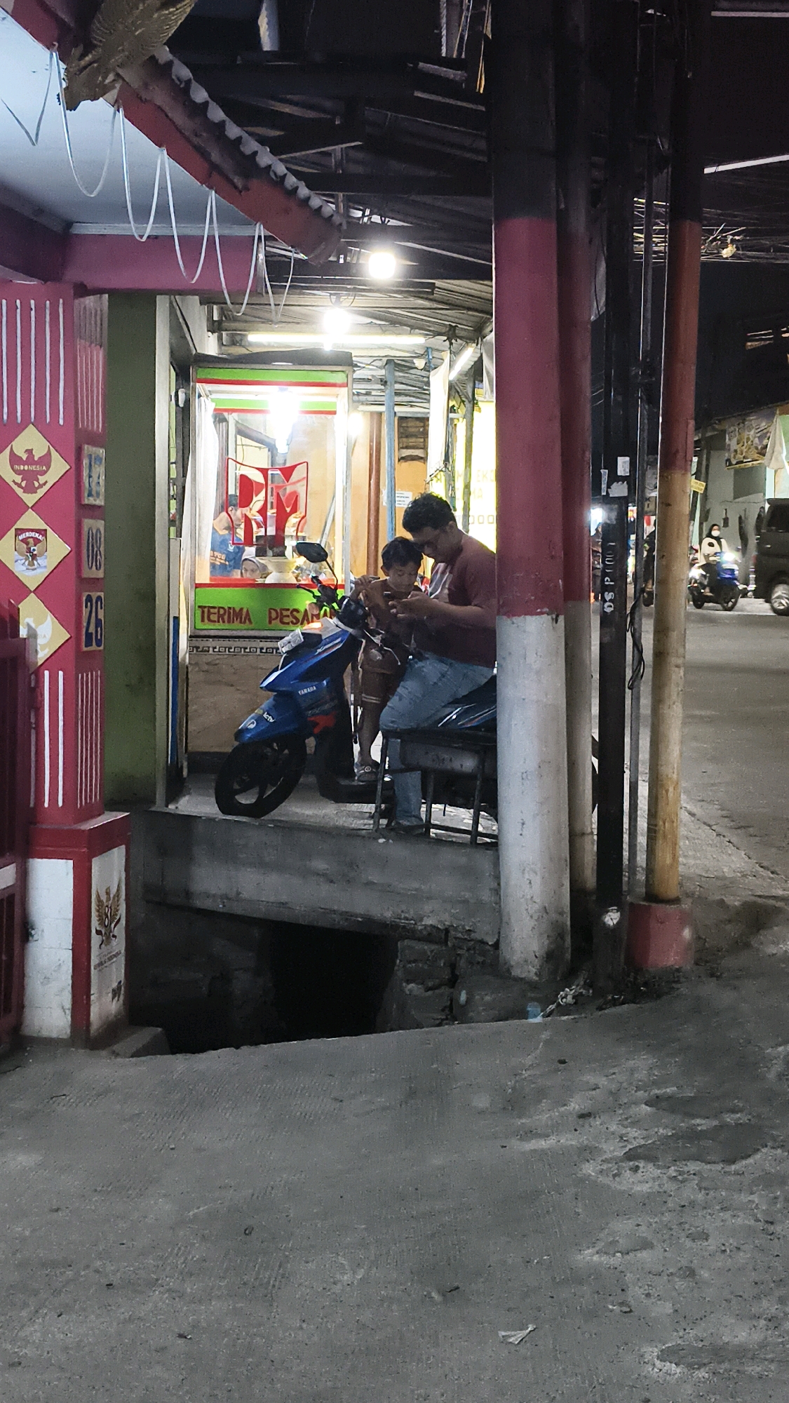 The image shows a street scene at night in Bekasi, Indonesia. A man and a child are seated on a motorcycle in front of a small establishment with a sign that reads "TERIMA PESANAN" (Take Orders). The establishment appears to be a food stall or small shop, with bright lights illuminating its interior. A blue Yamaha motorcycle is parked in front of them. The surrounding environment is dimly lit, with concrete pavement and several vertical poles. In the background, there are other people and vehicles, suggesting a moderately busy street. The scene is captured from a slightly low angle, looking up towards the establishment.