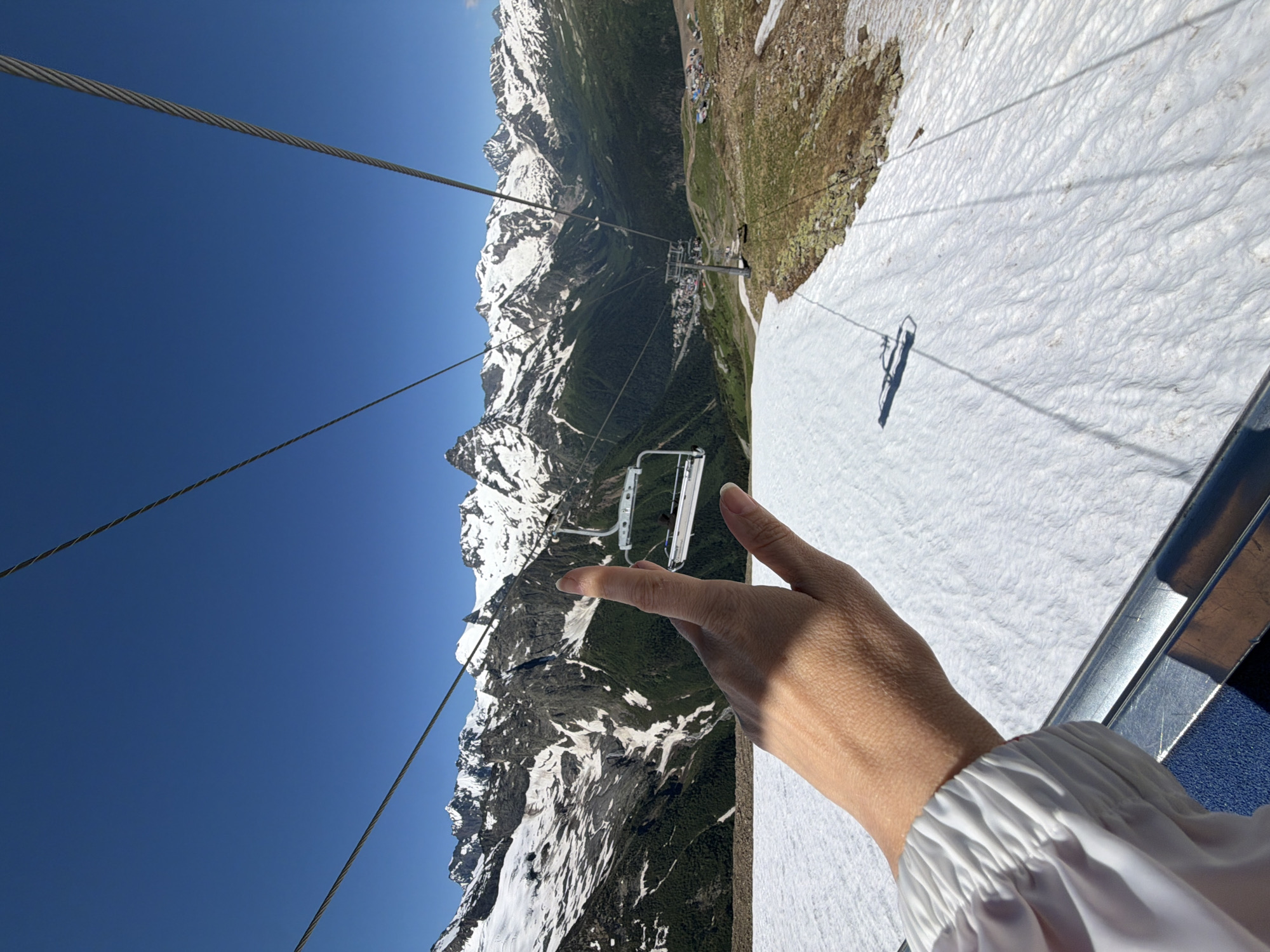 A person's hand is in the foreground, with their index finger pointing towards a ski lift in the distance. The ski lift consists of chairs suspended from cables, traversing a snowy mountain landscape under a clear blue sky. The mountains are snow-capped and rugged, with green slopes visible in the lower sections. The ski lift appears to be in operation, with several chairs visible. The hand is lightly tanned and has manicured nails. The person is wearing a white garment with ruffled sleeves. The scene suggests a sunny day, likely during the warmer months when snow persists at higher altitudes. The image evokes a sense of adventure and exploration in a mountainous environment.