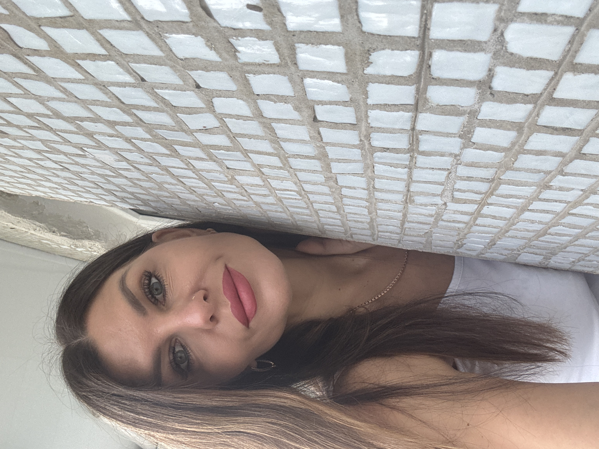 A woman with brown hair, fair skin, and red lipstick poses in front of a wall of white square tiles. She wears a white top and a delicate gold necklace. The location is Chaikovsky, Russia.