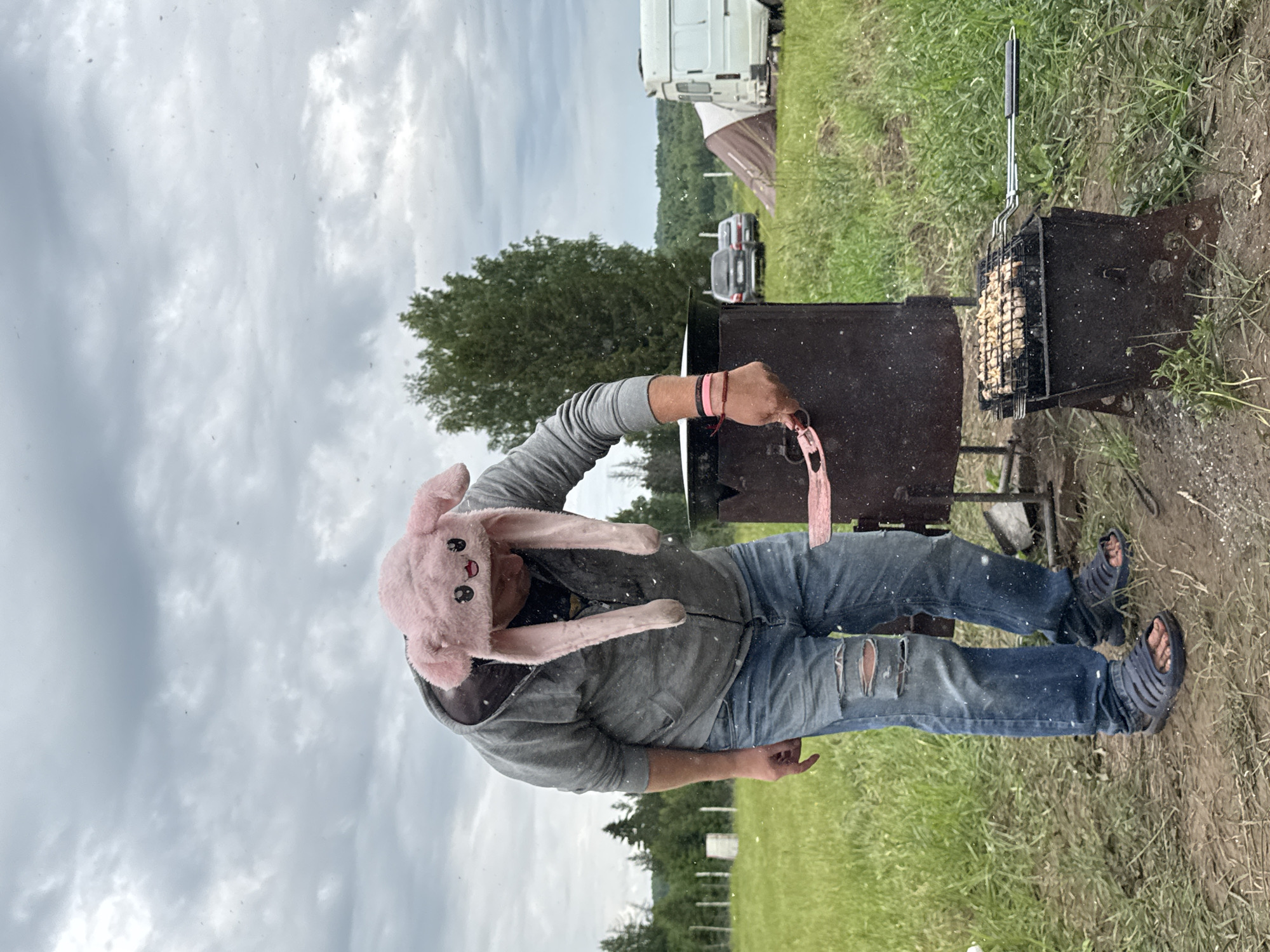 An outdoor scene captures a person grilling food under an overcast sky. The central figure, wearing a playful pink bunny hat, a grey hooded sweatshirt, ripped blue jeans, and dark sandals, stands next to a dark metal barbecue grill (mangal). They are holding pink tongs, appearing to turn or manage pieces of meat (possibly chicken) cooking on a metal grate over the coals. The person also has pink and dark wristbands on their right arm.
The background features a lush green grassy area, scattered trees, and hints of a rural or camping setting. Visible in the distance are a large white semi-truck, a dark car (possibly an SUV), and a tent. The sky is largely covered with light grey clouds, indicating an overcast day. Numerous small specks, possibly dust motes or light precipitation, are visible against the sky and throughout the image. The ground immediately around the grill is bare earth. The overall impression is of a casual outdoor cooking event. No visible text is present in the image. The location context is Большая Кивара, Russia.