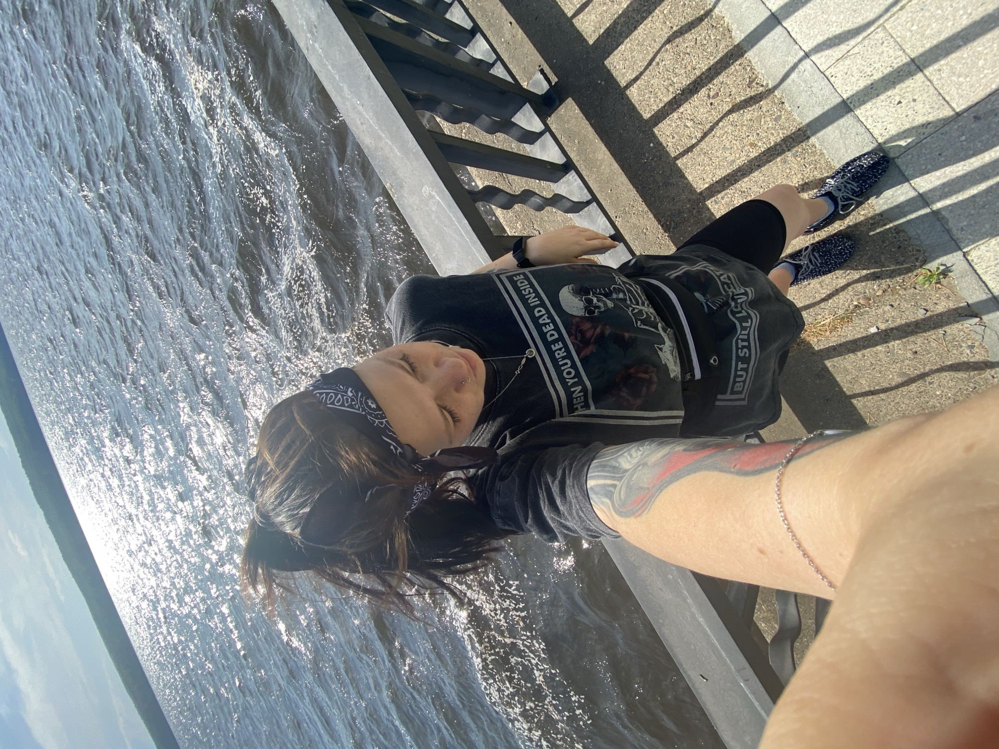 A medium shot depicts a woman taking a selfie with a body of water in the city of Chaikovsky, Russia, visible in the background. She is wearing a bandana on her head, a dark-colored t-shirt with graphic print on the front that partially reads "THEN YOU'RE DEAD INSIDE", dark bike shorts, and patterned shoes. The woman is positioned near a gray metal railing that runs along the waterfront, as indicated by the lapping water. The sidewalk area behind the railing is partially visible, showing patterns of sunlight and shadow cast by the railing itself. The woman's left arm, adorned with a tattoo and a silver bracelet, extends towards the camera, suggesting she's taking a selfie.