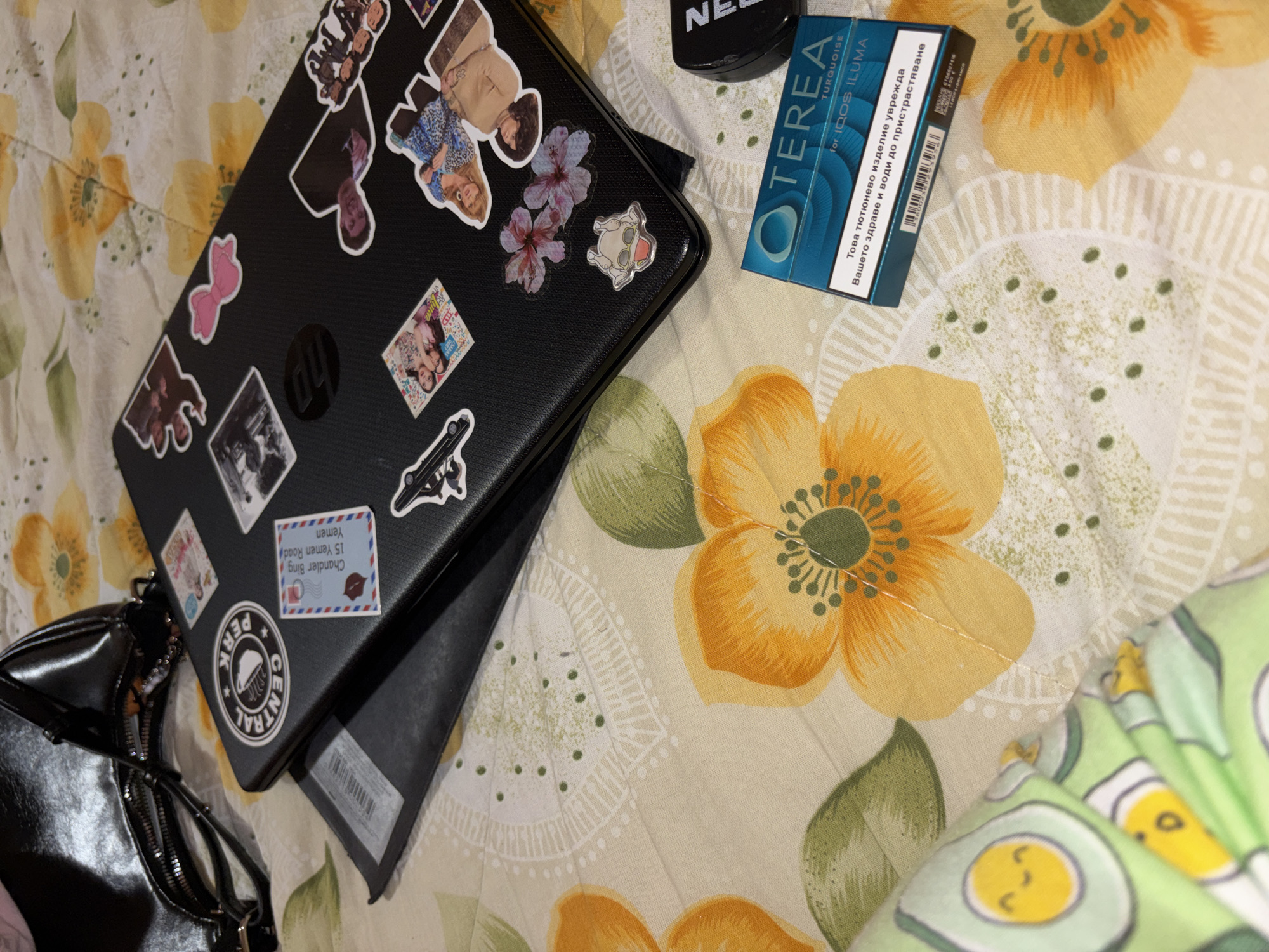 The image shows a close-up, slightly angled view of various objects resting on a floral-patterned fabric, likely a bedspread or similar textile. A black laptop, adorned with stickers of people and objects, is prominently positioned on the left side. Next to it is a black purse. Adjacent to the laptop is a pack of TEREA Turquoise for IQOS ILUMA. The sticker on the laptop shows Chandler Bing at 15 Yemen Road, Yemen.
The picture was not taken in the city of Garmen, Bulgaria, based on the stickers on the laptop indicating Yemen.
