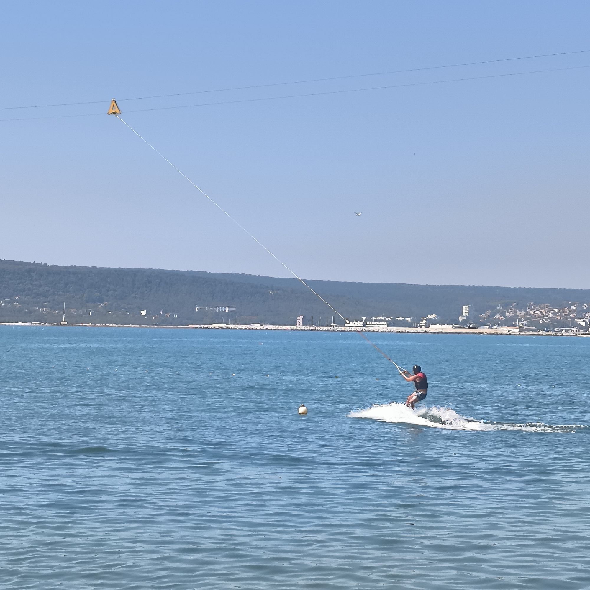 In Varna, Bulgaria, a person is waterskiing in the sea under a clear blue sky. The individual is wearing a life vest and holding onto a tow rope connected to a kite visible in the upper left quadrant of the image. The kite is orange and triangular in shape. The person is creating a white wake behind them as they move across the water. In the background, a coastline with lush green trees and buildings is visible, suggesting the location is near a populated area. The sea is calm with small ripples on its surface. A white buoy is also floating in the water to the left of the skier.