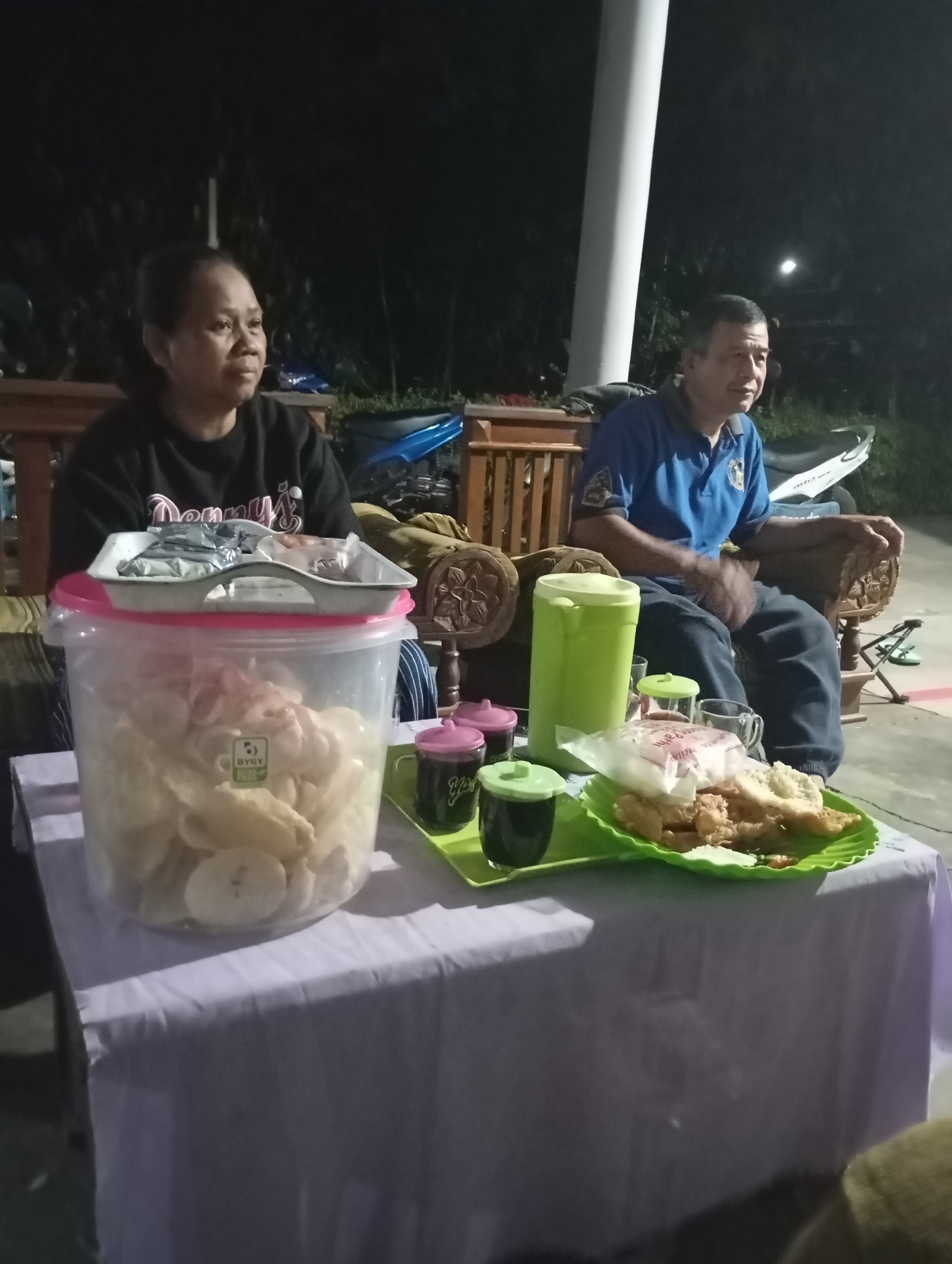 In the city of Wonogondo, Indonesia, an outdoor evening gathering is depicted. Two adults, a woman and a man, are seated on a wooden bench, facing the viewer. The woman, on the left, wears a black sweatshirt. The man, on the right, is dressed in a blue polo shirt and dark pants. In front of them, a table is laden with food and drinks. A large clear plastic container filled with snacks, likely crackers or chips, is prominently displayed. Adjacent to it are small containers of dark liquid, likely beverages, and a green tray holding fried food wrapped in paper. A green thermos is also present on the table. The background reveals a dimly lit outdoor setting with foliage and what appears to be a motorcycle. The scene suggests a casual social gathering.