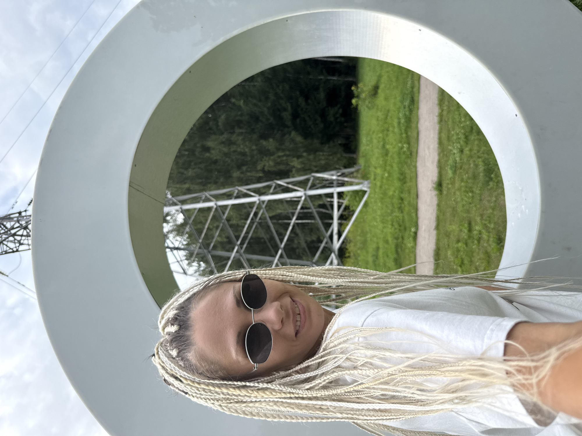 A young woman with blonde braided hair and sunglasses smiles at the camera. She is wearing a white t-shirt and appears to be standing in front of a large, circular, white sculpture. In the background, a transmission tower is visible amidst trees and a grassy field. The sky is cloudy. The image was taken in Rusanovo, Russia.