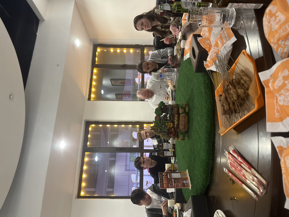 The scene depicts six individuals, five young adults and one middle-aged male, seated around a large, dark, circular dining table in a brightly lit indoor restaurant in Huangdao District, China. Overhead lighting and decorative string lights outlining two large windows illuminate the space, revealing blurred external reflections.
A central green circular mat on the table supports a multi-tiered decorative structure resembling an ornate miniature building or landscape feature. Food items include numerous skewers of grilled meat on an orange tray, various dishes on individual plates, and packaged chopsticks. Beverages consist of beer bottles, including one identified as Tsingtao Pure Draft, and water bottles, alongside empty and full clear drinking glasses. A black tissue box and a stand-up promotional card with Chinese characters, including "扫码支付" (scan code to pay), are also on the table. Small crumpled orange-and-white paper wrappers are scattered on the table surface.
The individuals are casually dressed. The person on the far right, wearing a plaid shirt, is actively eating with chopsticks. The middle-aged male in a white shirt is looking down at his plate. Other individuals are generally observing the table or their food, consistent with a group meal. No direct interpersonal interactions are distinctly captured.