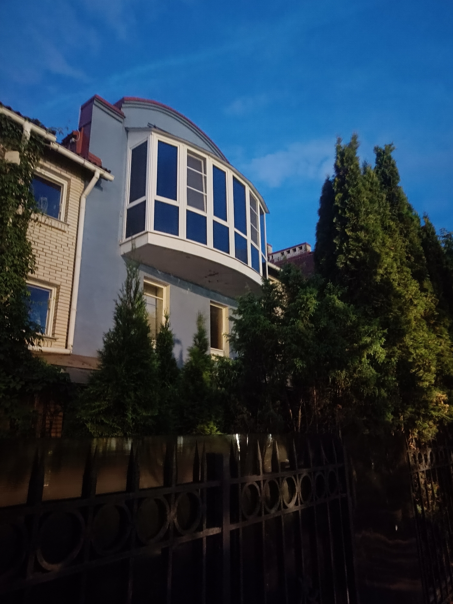 The image depicts a residential building with a distinctive curved bay window, situated at dusk or twilight. The building features a lighter colored brick facade on the left, juxtaposed with a smooth, pale blue wall on the right. A dark, ornate metal fence is prominently displayed in the foreground, partially obscuring the view of the building's lower section and the surrounding vegetation. Tall evergreen trees, likely conifers, flank the right side of the building, their dense foliage contrasting with the clear, deep blue sky above. The architectural style and the surrounding greenery suggest a potentially affluent residential area. Without specific landmarks, precise localization within Saint Petersburg, Russia is not possible.