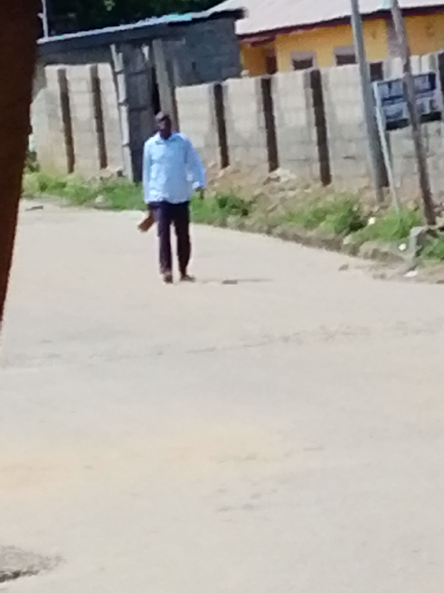 A man is walking along a paved road in Lokoja, Nigeria. He is wearing a light blue long-sleeved shirt and dark trousers, and he is carrying an object in his hand. To his right is a concrete block fence with dark vertical accents, along with a signpost and some vegetation. Behind the fence, there are buildings with corrugated metal roofs, one painted yellow. The road surface is light-colored, and there is foliage visible in the background. The angle appears to be from a slightly elevated vantage point, suggesting the photo was taken from a window or another high location.