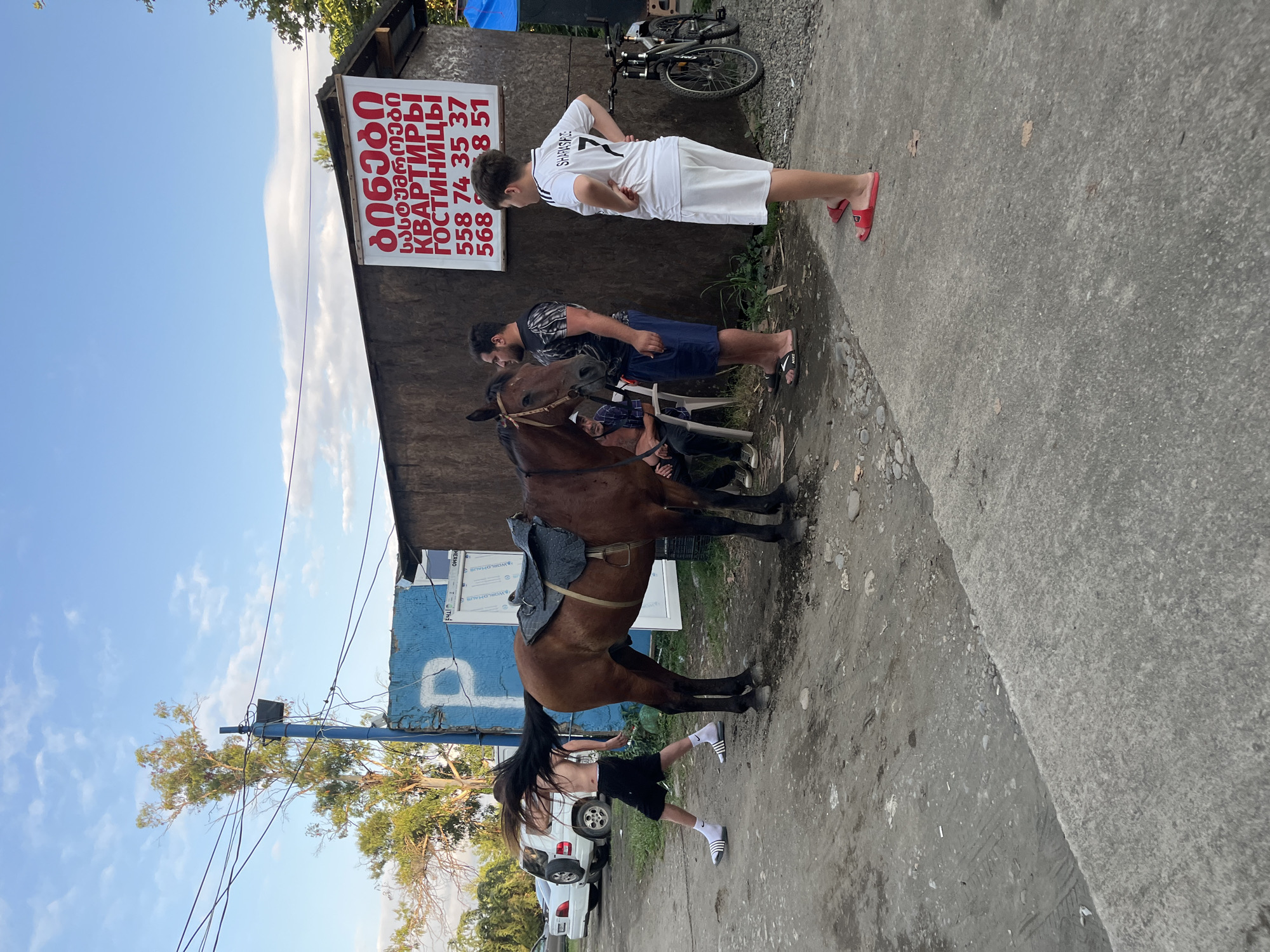 In Ureki, Georgia, a brown horse stands tethered near a small wooden building. A saddle is placed on its back. A young person with long, dark hair, wearing black shorts and white athletic slides, is seen in motion near the horse's rear, their hair dramatically flowing. Two men stand to the right of the horse, one in a black t-shirt and shorts, the other in a white t-shirt with the number 7 and white shorts. A younger person in a white t-shirt and shorts with red slides is also present. An older man sits on a chair to the left. A bicycle is parked to the far right. The scene is set on a dirt and paved ground surface with a blue structure bearing a white 'P' visible in the background. A red and white sign is affixed to the wooden building, displaying text in Georgian and Russian characters, along with phone numbers. The sky is partly cloudy.