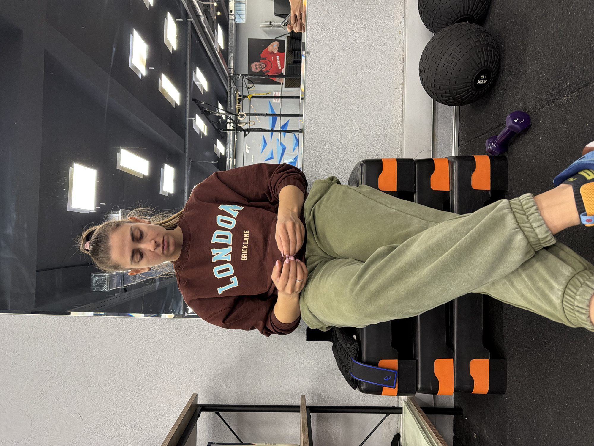A young woman with light skin and blonde hair pulled back in a ponytail sits on a black and orange plyometric box in what appears to be a gymnasium. She is wearing a brown sweatshirt with "LONDON" printed vertically on the left and "BRICK LANE" printed horizontally underneath. She also wears green sweatpants. The gym has exposed pipes and many square lights on the ceiling. In the background is a pull-up bar rig and a poster of a man in a red shirt. Next to her on the floor are two black medicine balls and a small purple dumbbell.
Determining the exact location in Sofia, Bulgaria, based solely on the image is not possible.