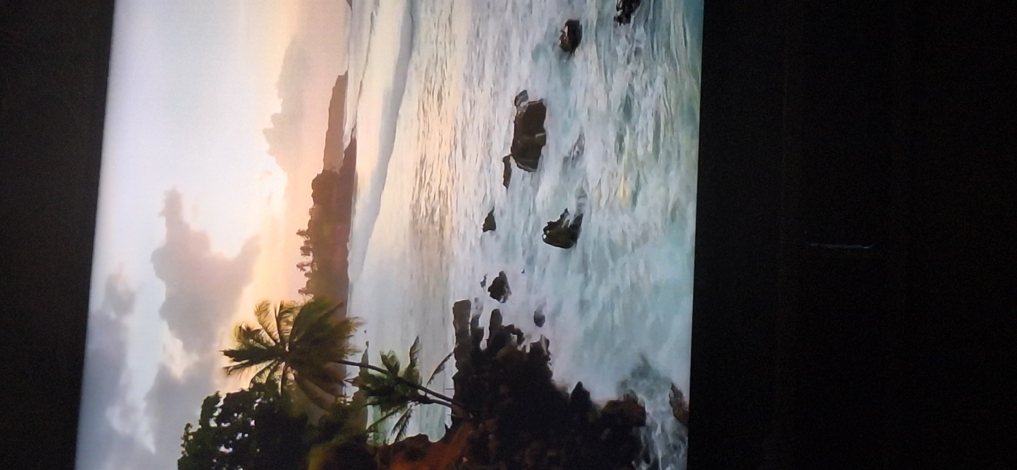 The image shows a tropical beach scene at sunset. Palm trees and lush vegetation line the shore, which is composed of dark rocks and sand. Waves crash against the rocks, creating white foam. The sky is filled with dramatic clouds tinged with orange and pink from the setting sun. The ocean stretches out to the horizon, with gentle waves rolling in. The overall impression is one of natural beauty and tranquility. There is no indication of this scene being in Bila Tserkva, Ukraine.