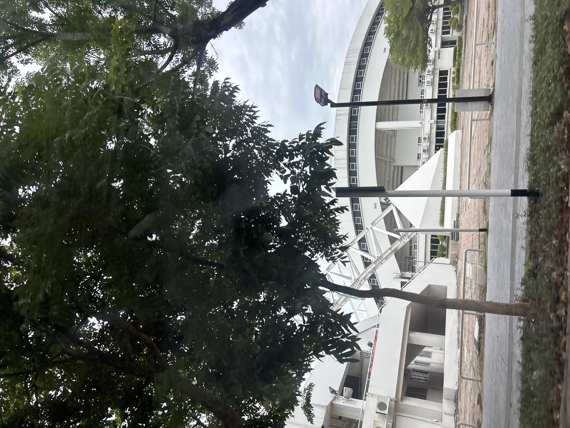 The image depicts an outdoor scene on an overcast day, featuring a large, modern white building with a curved roofline, possibly a stadium or convention center. The building's facade has a repeating pattern of windows and air conditioning units. In the foreground, several trees with dark green foliage partially obscure the view of the building. The ground in front of the building is paved with bricks and has metal railings. A street lamp with a distinct modern design stands near the building. The environment appears to be urban, likely in Tha Khlong Town Municipality, Thailand, given the context. There are no visible people or animals in the image. The overall atmosphere is neutral and the lighting suggests diffused daylight.