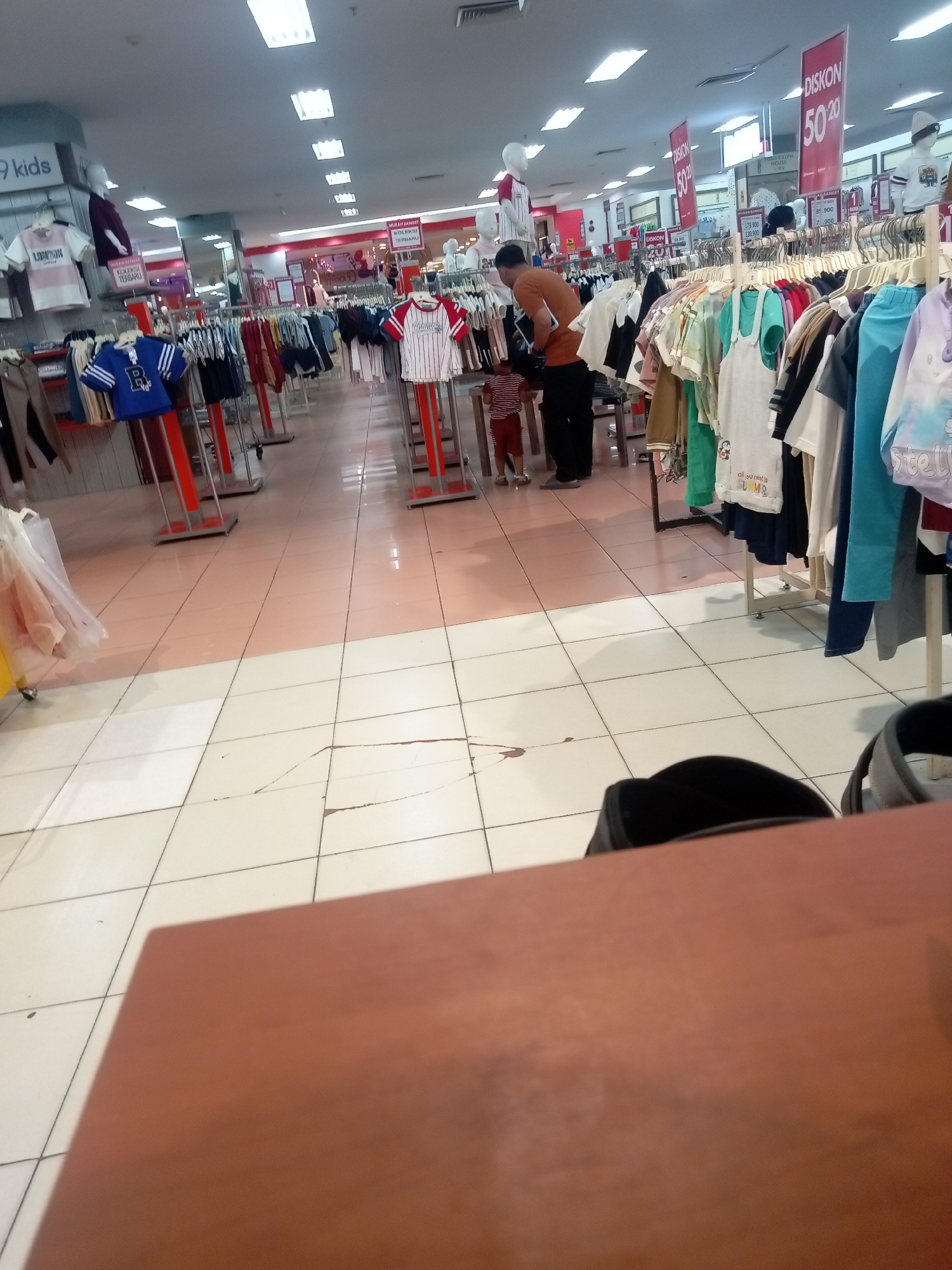 The image depicts the interior of a retail store, likely a clothing department in a department store or supermarket, in Batam, Indonesia. The scene is brightly lit with fluorescent ceiling lights. Racks of children's clothing are displayed on both sides of an aisle, with some adult clothing also visible. Several red promotional banners with "DISKON 50%" are prominently displayed, indicating a sale. In the center of the frame, a person in an orange shirt and dark pants is interacting with a child wearing a striped shirt and shorts. The floor is tiled with a pattern of light and darker tiles. The overall impression is that of a busy retail environment.