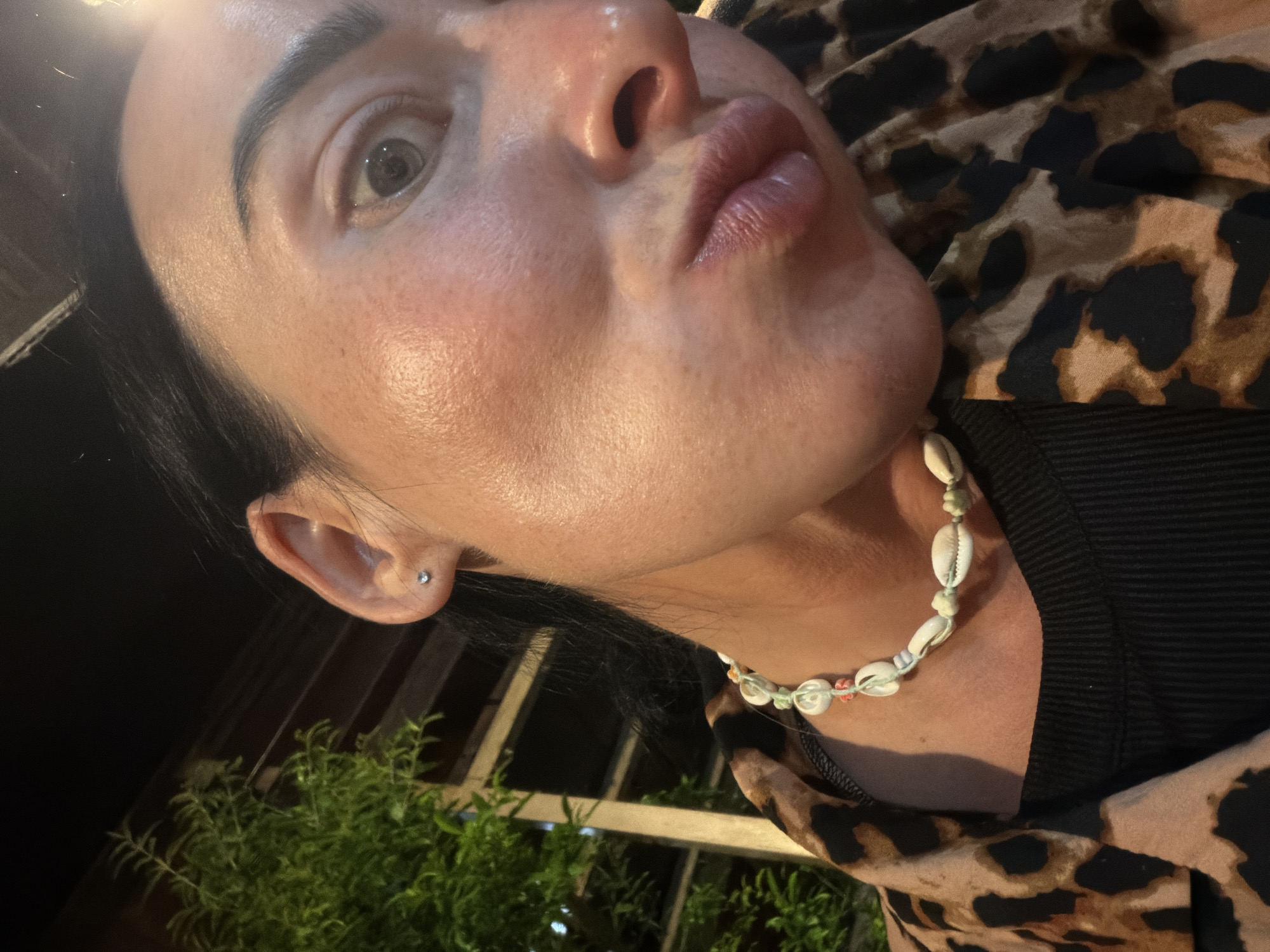 A close-up shot captures a woman's face from the chin up, with her head tilted. She has dark hair pulled back, revealing her right ear, adorned with a small stud earring. Her skin appears slightly flushed, with visible pores and some freckles. Her right eye is open and looking towards the camera, while her lips are puckered. She is wearing a black ribbed top and a leopard-print jacket. A necklace made of shells and beads rests on her collarbone. In the background, out of focus, is lush green foliage and what appears to be part of a building structure with vertical beams. The lighting suggests an outdoor setting at night, possibly on a balcony or patio. The location is specified as Sochi, Russia.