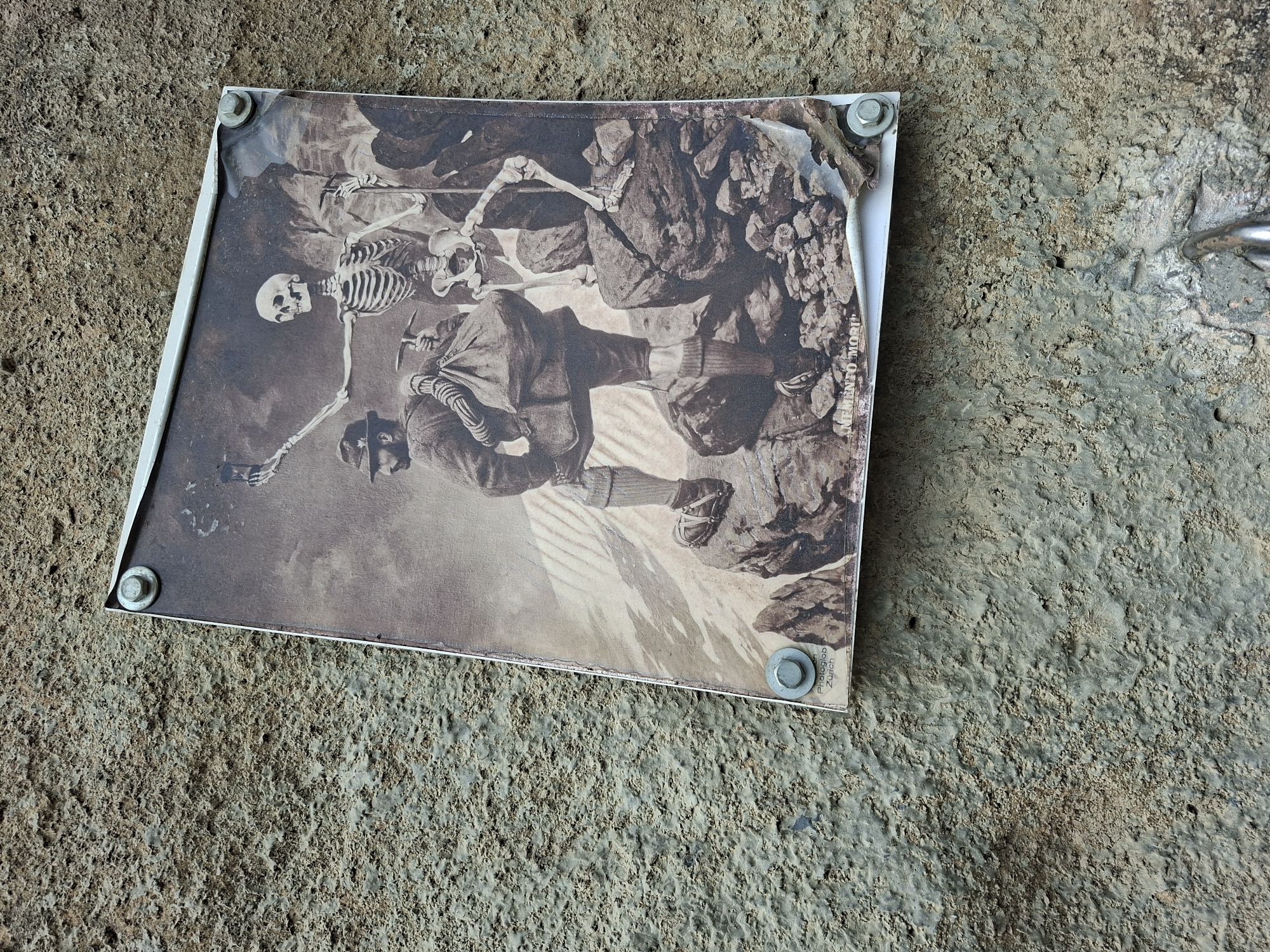 A sepia-toned print, depicting two skeletons and a human in mountaineering attire, is affixed to a textured concrete wall. The print shows one skeleton holding a camera aloft, as if taking a selfie, while another skeleton stands beside it. A man in a hat and mountaineering gear, carrying a large bag, is positioned in the foreground. The print is secured to the wall by four metal screws, one of which is visible in the bottom right corner, partially obscuring the text "MEMENTO MORI" at the bottom of the print. The wall appears weathered and the print itself shows signs of damage and peeling. The overall scene suggests an outdoor setting, possibly a tourist trail or viewpoint, in Děčín, Czechia.