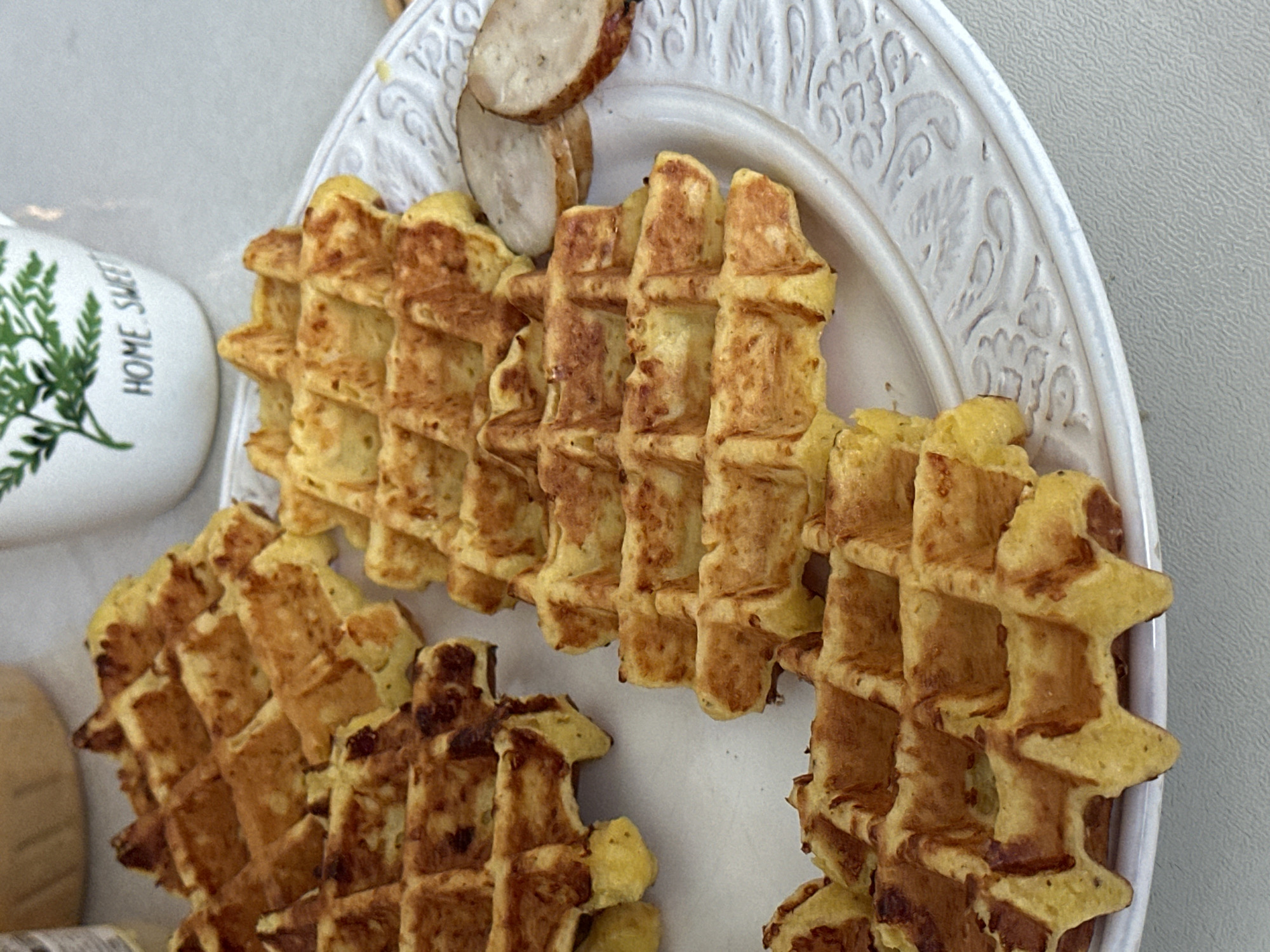 A close-up, overhead shot displays several golden-brown waffles arranged on a white, embossed plate. The waffles, cooked to a crisp texture, are slightly charred in some areas. To the right of the waffles, two slices of what appear to be sausage or a similar cured meat are visible. In the background, a white mug with a green fern design and the words "HOME SWEET" is partially visible. The setting appears to be a table, possibly in a kitchen or dining area, with a light-colored surface. No specific location in the city of Рабочий, Russia, can be determined from the visual information provided.