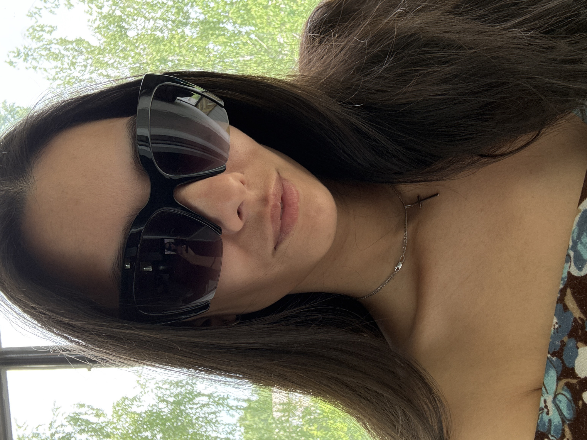A woman with long, dark hair and sunglasses is posing for a photo. She is wearing a silver necklace with a cross pendant and a floral-patterned top. The background shows green foliage and a window. The location is not definitively identifiable as Chaikovsky, Russia from the given image.