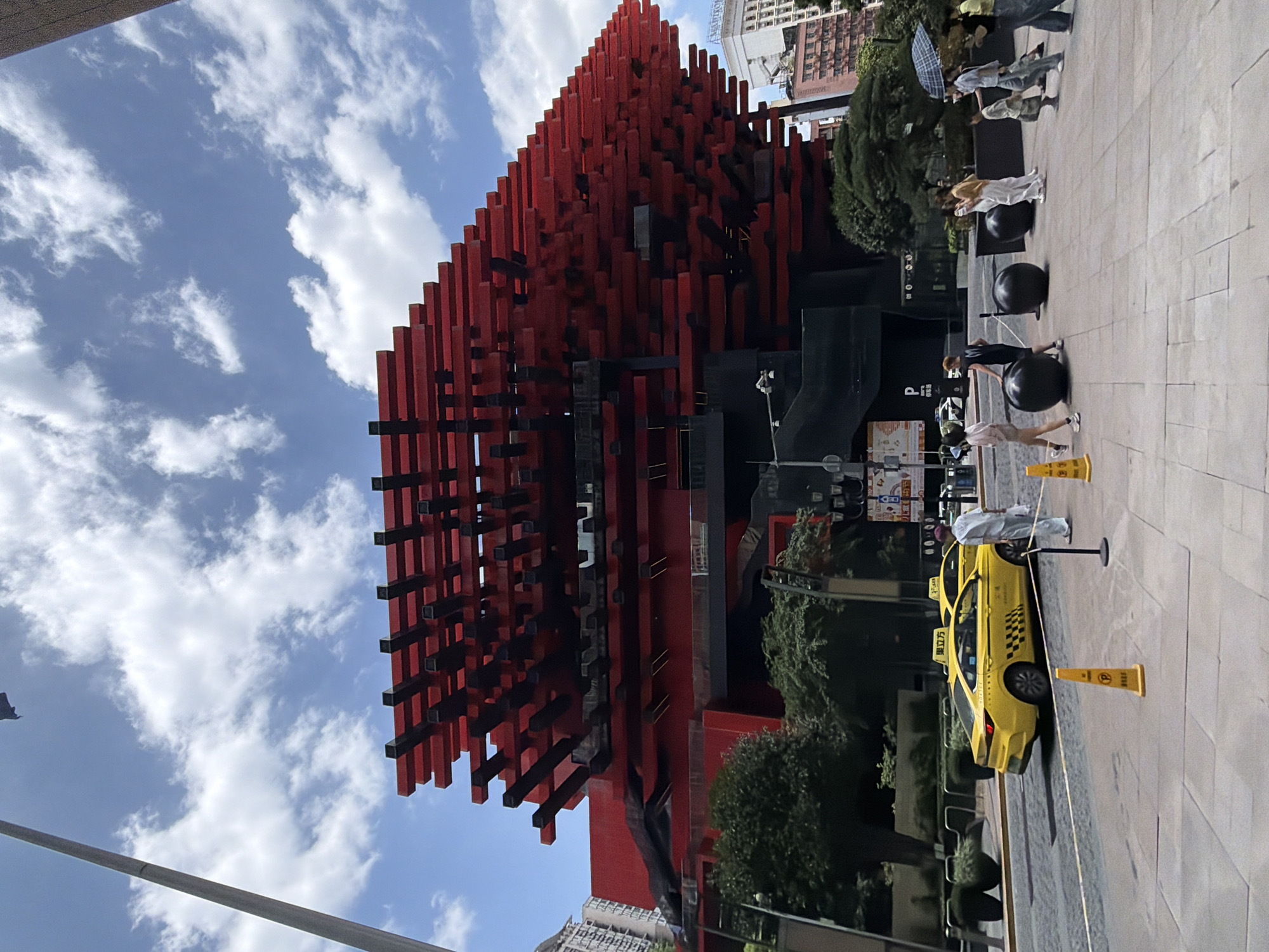 The image shows a wide-angle view of a modern building with a striking red and black facade, located in the Yuzhong District of China. The building's upper portion is covered in a layered, cantilevered structure made of red horizontal beams supported by black vertical elements. Below this, the building is predominantly red with black sections.
In the foreground, a street scene unfolds on a paved pedestrian area. A yellow taxi is parked on the left side of the road, with another yellow taxi partially visible behind it. Several people are walking on the pedestrian area. On the right, a group of individuals is walking, some with umbrellas. Two large black spherical sculptures are placed on the pedestrian area between the building and the sidewalk.
The environment is characterized by a bright, cloudy sky and lush green trees planted along the building's base. Other buildings are visible in the background, suggesting an urban setting. The overall impression is of a contemporary urban landscape with a focus on architectural design.