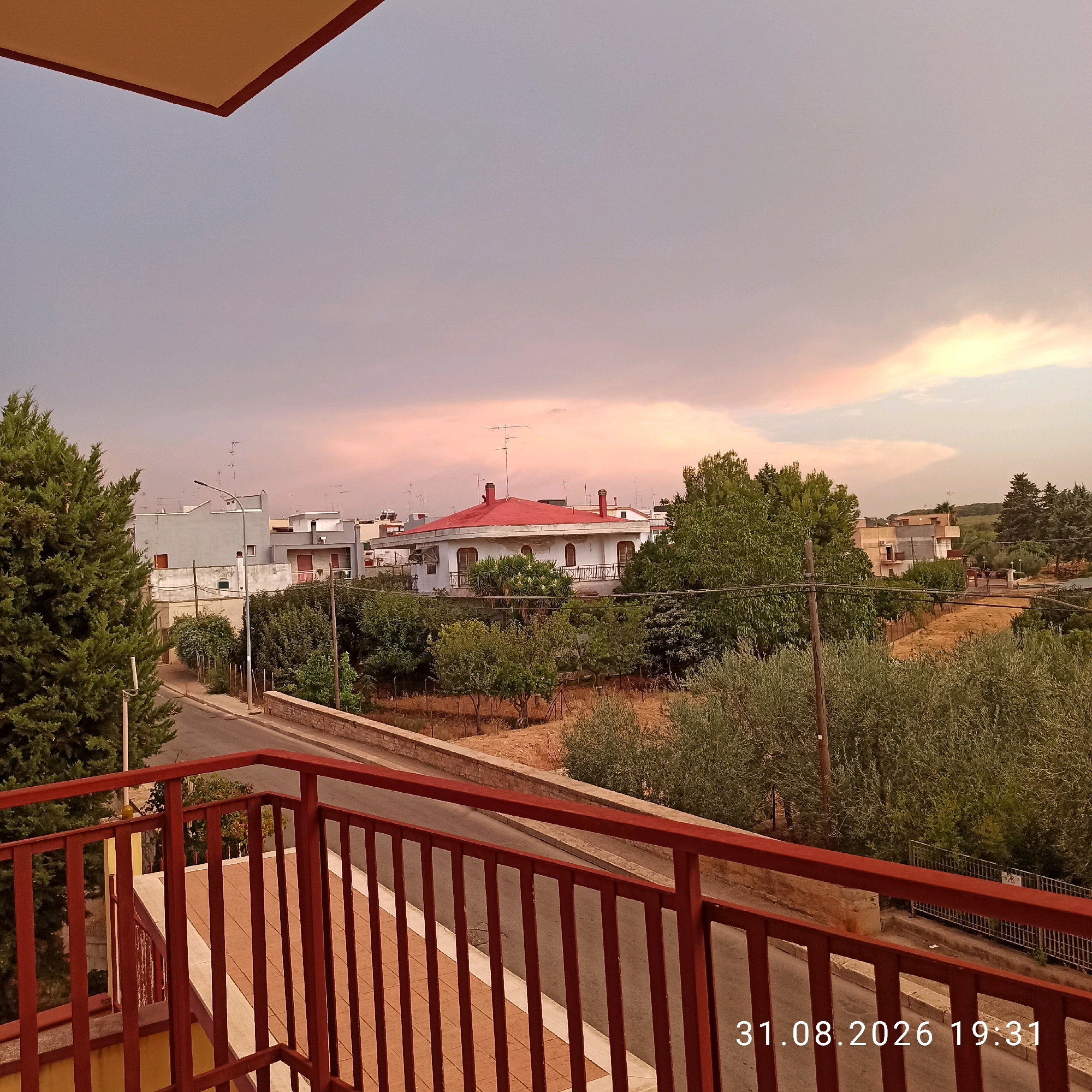 A low-angle, full shot captures an outdoor scene at dusk from a balcony. The foreground features a red metal railing. Beyond the railing, a street curves to the left, bordered by trees and a stone wall on the right. In the midground, several multi-story buildings are visible, some with red-tiled roofs. A prominent building with a red roof and arches on its balcony stands out. The background displays a hazy sky with subtle pink and orange hues, indicative of sunset. The timestamp "31.08.2026 19:31" is overlaid on the bottom right of the image. The overall setting appears to be a residential area in Bitonto, Italy, with a mix of vegetation and urban structures under a twilight sky.