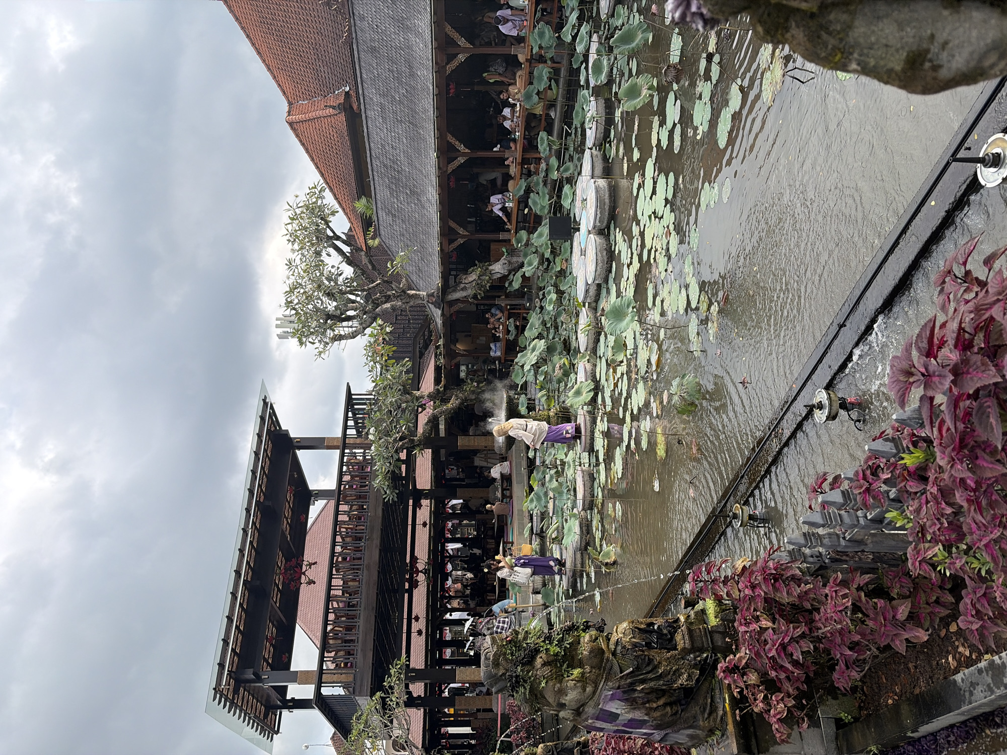 This is an outdoor scene in Ubud District, Indonesia, featuring a lotus pond with stepping stones and several people. In the foreground, a stone statue covered in moss and vines stands to the left. To the right of the pond, a second, less moss-covered stone statue is visible. People are scattered throughout the scene, with some standing on the stepping stones in the pond and others seated at tables in an open-air restaurant with a multi-level wooden structure. The restaurant has a red-tiled roof and a glass-covered upper level. The sky is overcast.