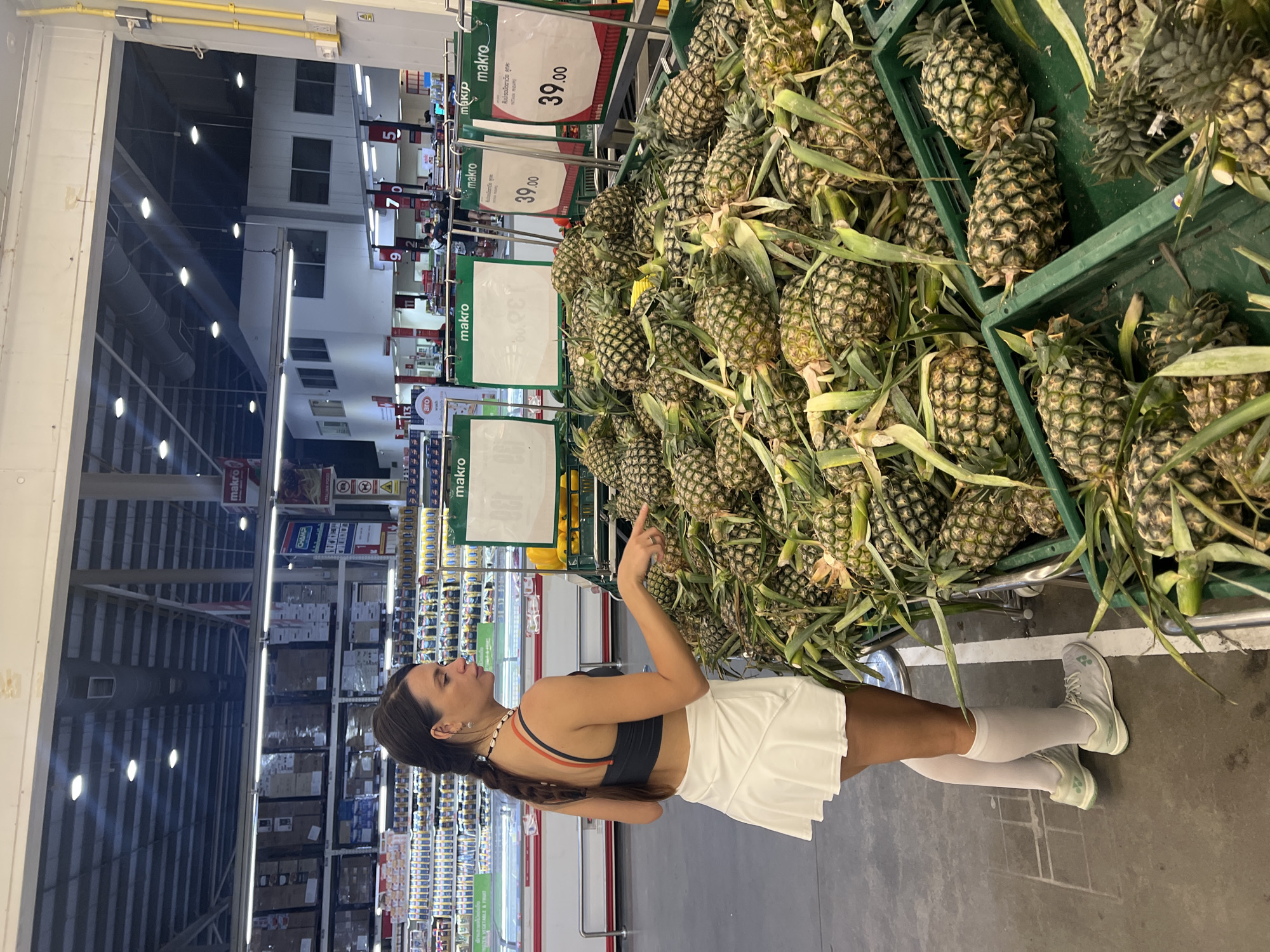 The image shows a woman pointing at pineapples in a Makro supermarket. She is wearing a dark sports bra, a white tennis skirt, and white knee socks with light green sneakers. There are many pineapples in green plastic crates, with price tags that indicate the price is 39.00 Thai Baht. Shelves with various items are visible in the background. The store is located in Baan Bo Phut, Thailand.