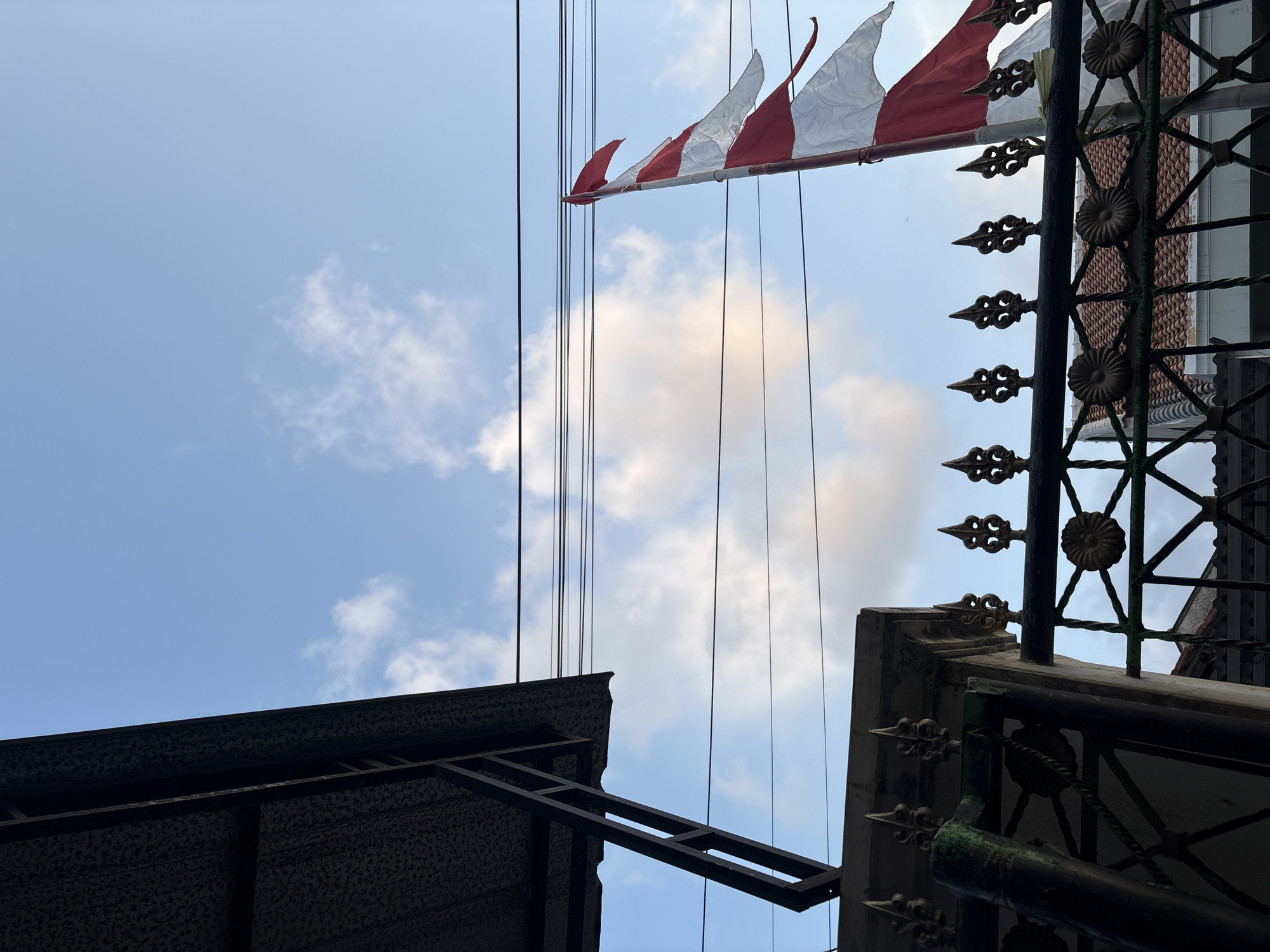 An Indonesian flag, comprised of red and white horizontal stripes, flutters on a pole against a bright blue sky dotted with white clouds. The flag is positioned to the right of the frame, partially obscured by the overcast sky. In the foreground, a black metal fence with ornate finials runs horizontally across the lower portion of the image. The fence is partially obscured by shadows cast by the overhead structure of a building, which occupies the upper left quadrant of the image. The building appears to be a covered walkway or awning, supported by dark metal beams. The scene is likely set in Palimanan, Indonesia, suggested by the context of the Indonesian flag. The overall atmosphere is one of a bright, possibly breezy day.
