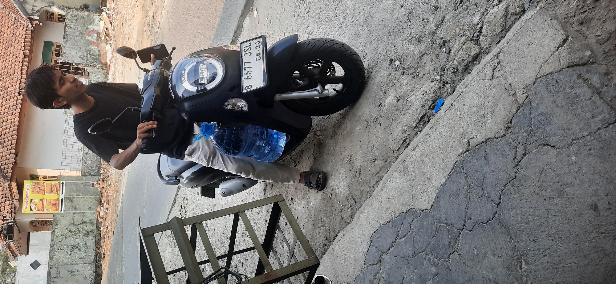 A young man wearing a black t-shirt and light-colored pants sits on a black scooter on a cracked concrete surface. He is holding the handlebars and looking down. A large blue plastic water bottle is strapped to the side of the scooter. In the background, a building with a tiled roof and a sign that reads "Risol Yummy" is visible. A metal rack stands to the left of the scooter. The scene appears to be outdoors in a street or roadside setting in Cisauk, Indonesia.