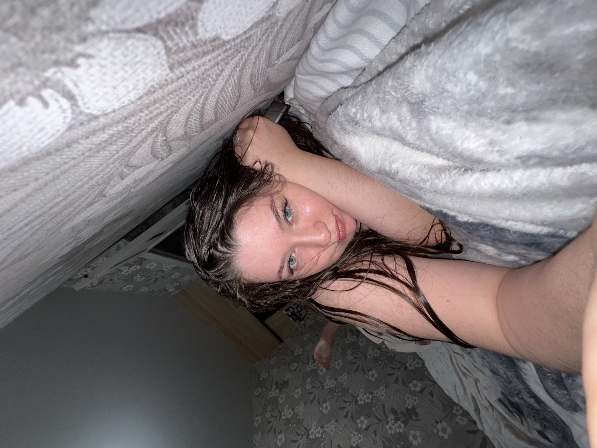 This media file captures a young woman from a low-angle, head-on perspective, appearing to be lying in bed. She has dark, wet-looking hair and light-colored eyes, looking directly at the camera with a neutral to slightly smiling expression. Her bare arm, showing some hair, is bent with her hand near her head, clutching a dark electronic device, likely a phone.
She is surrounded by various bedding textures: a light grey patterned fabric on the left side, and a fluffy, light grey blanket partially covering her on the right. A striped sheet or blanket is also visible near her head. In the background, a wooden furniture surface is partially visible on the left, and a grey and white floral or leaf-patterned wallpaper adorns the wall, near her visible bare foot.
The scene is indoors, likely a bedroom, with dim, artificial lighting suggesting it could be evening or night. The overall atmosphere is relaxed and intimate. The location context for this media file is Гамово, Russia. No visible text is present in the image.