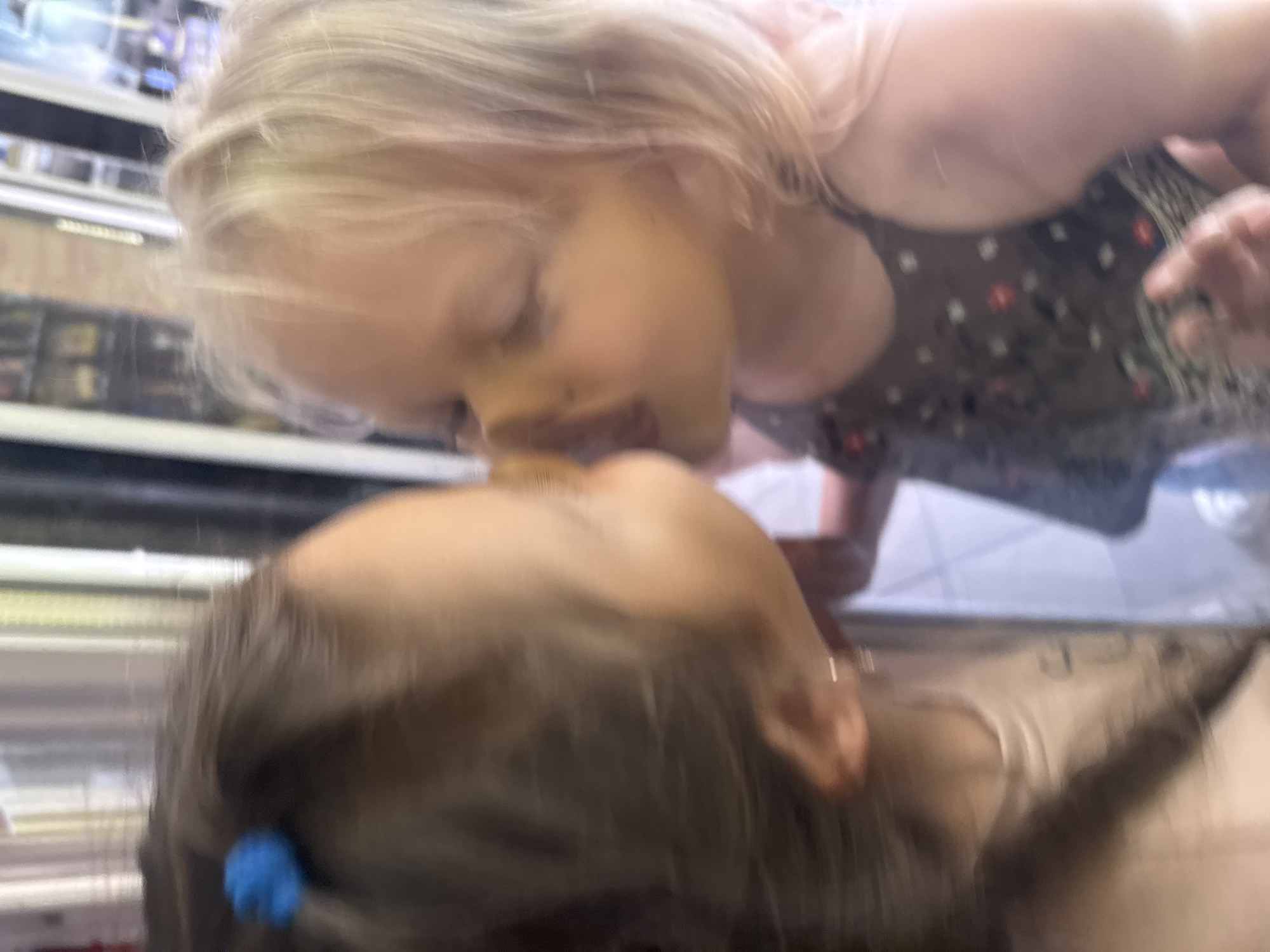 The image shows a close-up, blurred view of two young children interacting. The child on the left has dark hair pulled back with a blue hair tie. The child on the right has blonde hair and is wearing a dark sleeveless dress with a floral pattern. The children appear to be very close, possibly sharing a kiss or hug, with their faces touching. The background is out of focus but suggests an indoor environment with vertical elements that could be shelving or part of a structure. The overall impression is a moment of affection captured in a casual setting. The location is stated as Yalta, Ukraine.