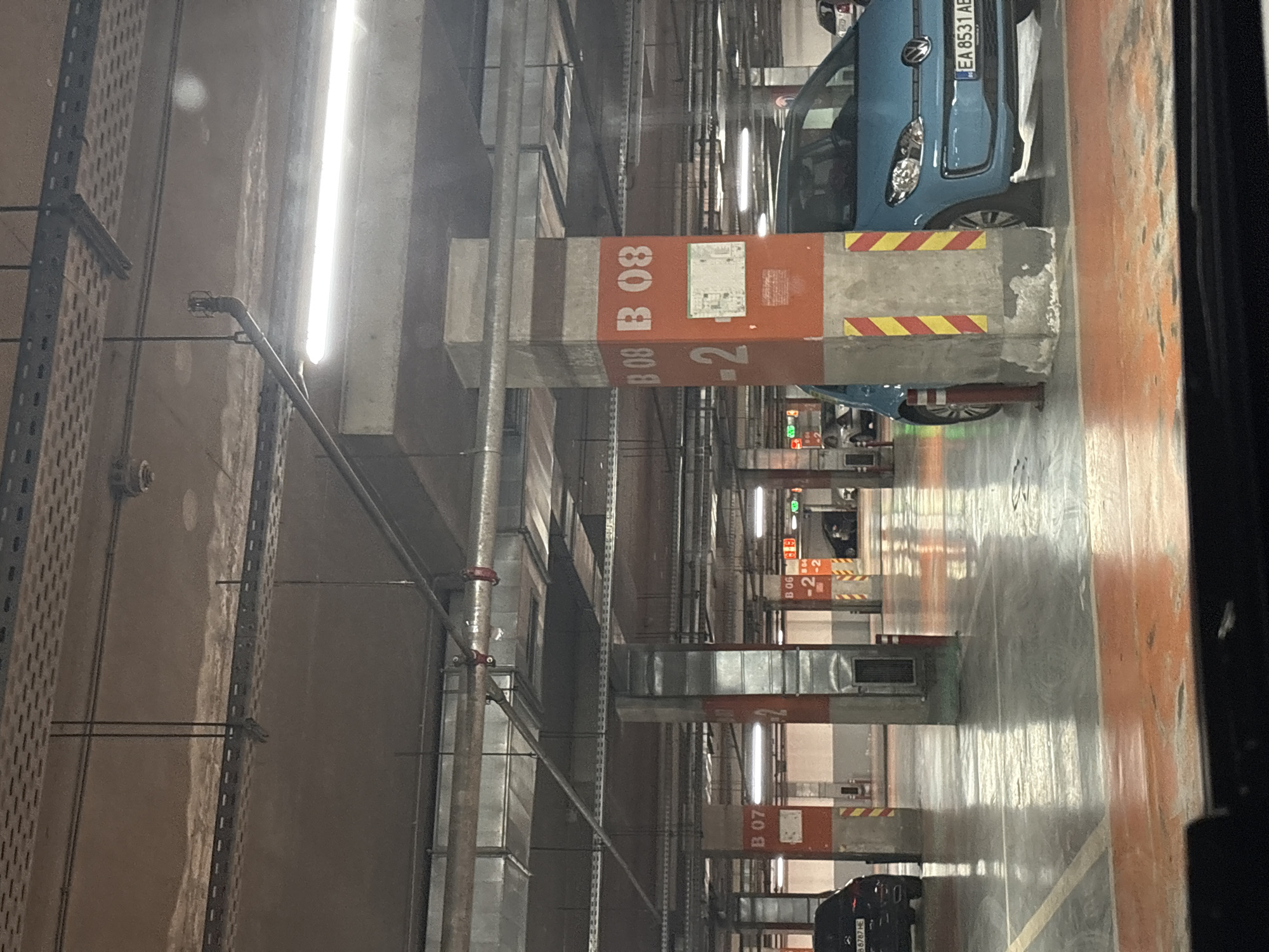 Here is a detailed description of the image:
The image shows the interior of an underground parking garage. The scene is lit by long fluorescent lights that run along the ceiling. The parking garage is divided into sections labeled "B 06," "B 07," and "B 08" with prominent orange and white numbers painted on the support pillars. Yellow and red stripes adorn the upper portion of some of the pillars. Cars are parked in the garage, including a blue Volkswagen with a license plate reading "EA 8531 AE" and a dark vehicle with a license plate reading "E 88787 HE." Various pipes, ventilation ducts, and metal supports are visible along the ceiling and walls. The garage has painted lines defining the parking spaces. The scene appears to be in an urban setting, likely in Sofia, Bulgaria, given the vehicle registration plates.