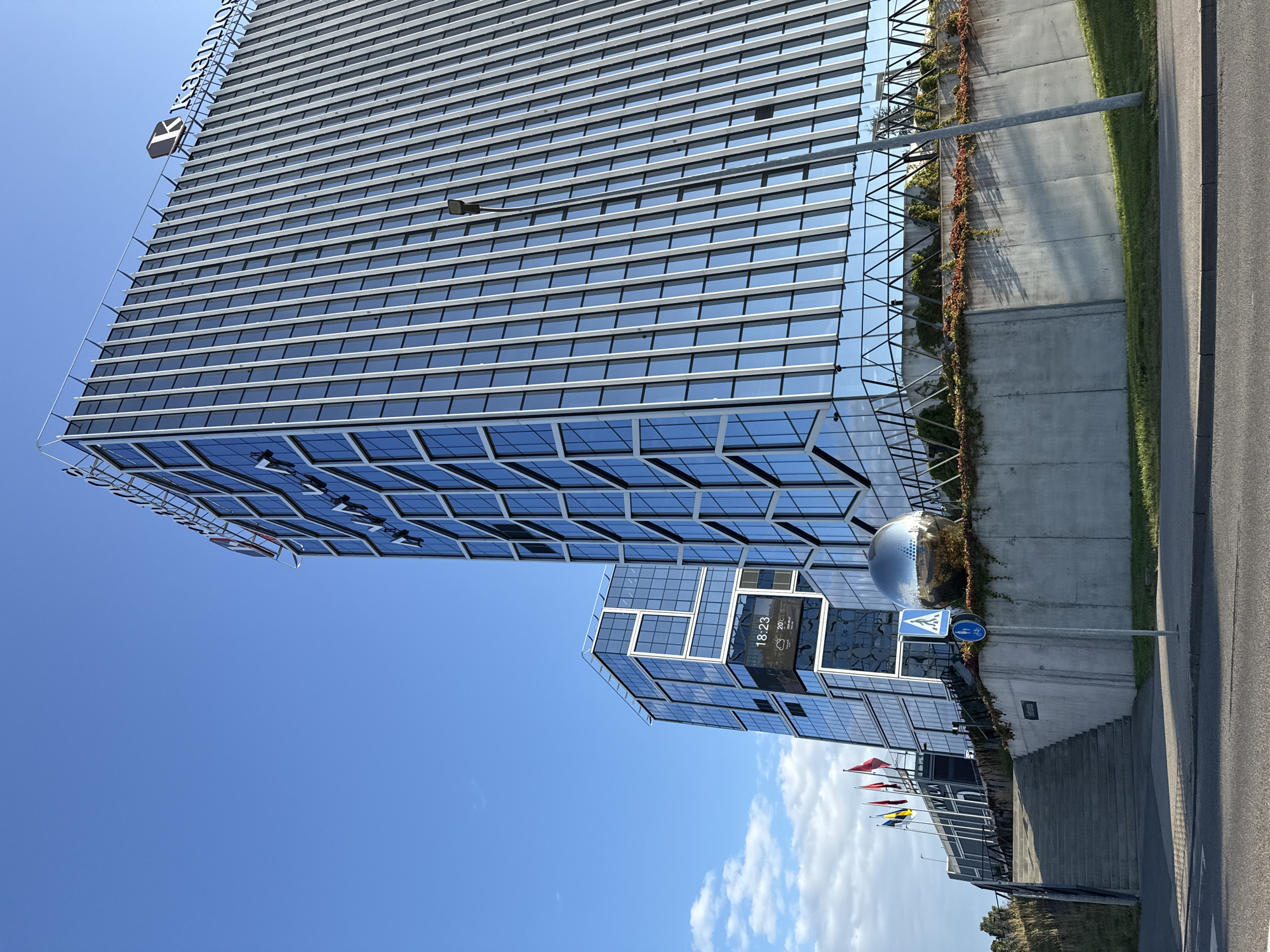The image depicts a modern architectural complex featuring two buildings with glass facades against a clear blue sky. The taller building, on the right, has a predominantly gray and glass exterior with vertical white accents. The smaller building on the left is also glass-fronted, with a more complex, segmented design. A large, reflective spherical sculpture is situated in front of the smaller building, on a raised concrete embankment adorned with some greenery. To the left of the buildings, a set of concrete stairs leads up to an elevated walkway. Several flags are visible on flagpoles near the stairs. In the foreground, a street runs horizontally across the bottom of the image, with a pavement and a grassy area. Traffic signs, including a pedestrian crossing sign and a bicycle path sign, are also present near the concrete embankment. The image is taken from a low angle, emphasizing the scale of the buildings. The scene appears to be a business or commercial district, likely in an urban setting. Based on the "Kaanos" logo on the taller building and the style of modern architecture, this is likely the Kaanos Business Centre in Tallinn, Estonia.