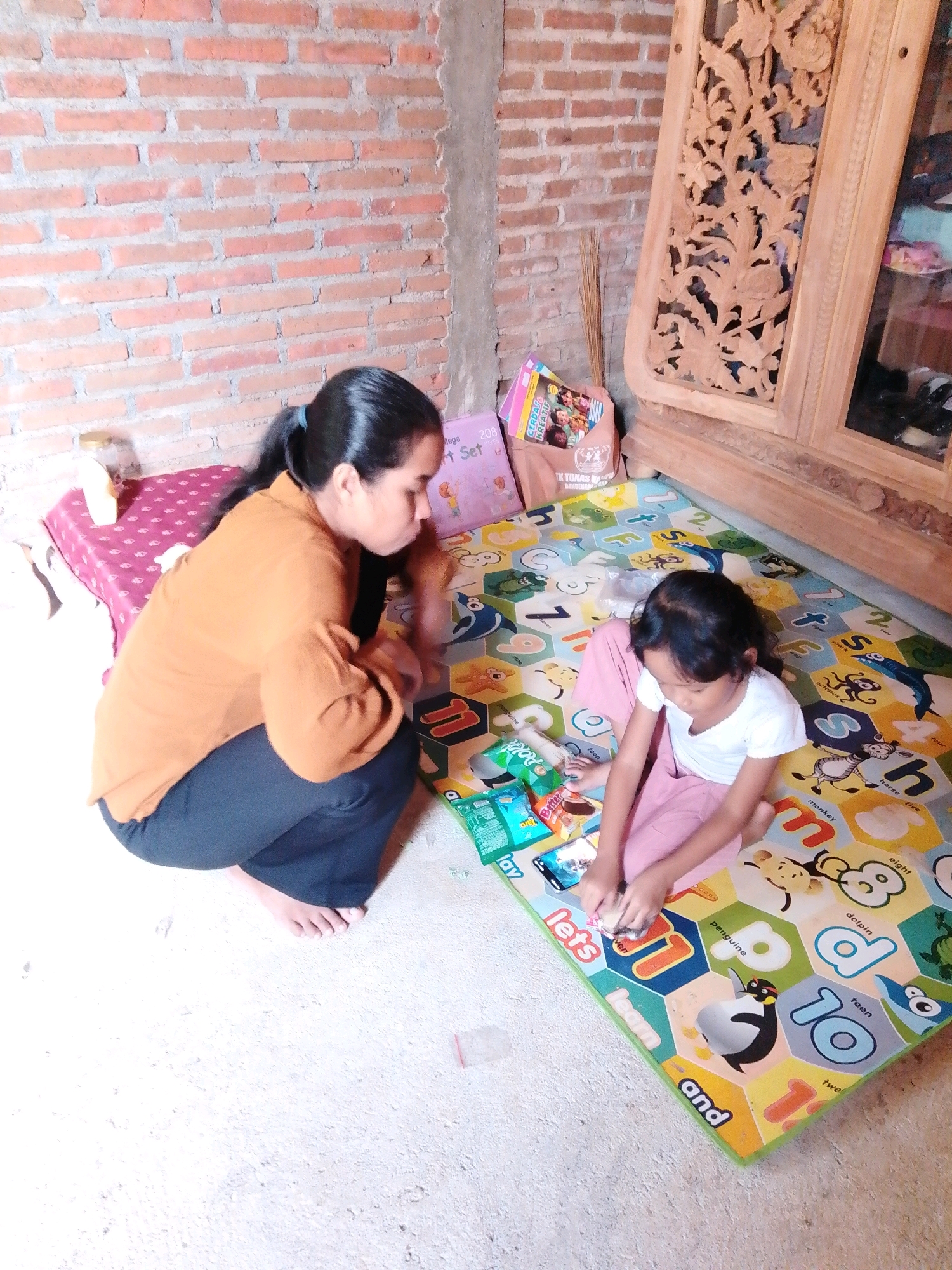 In the city of Cikal, Indonesia, an Indonesian woman in her 30s, wearing an orange long-sleeved shirt and black pants, is squatting on a patterned mat. She is interacting with a young Indonesian girl, approximately 6 years old, who is wearing a white t-shirt and pink pants. The girl is seated on the mat and appears to be playing with snacks and a mobile phone. The mat has colorful hexagonal patterns with letters and animal illustrations. Several snack packets are scattered around the girl. Behind them, a brick wall forms the backdrop, and a wooden cabinet with intricate carvings stands to the right. A broom is leaning against the wall near the cabinet. The floor is concrete. The scene suggests a domestic setting where the woman and child are engaged in a shared activity.