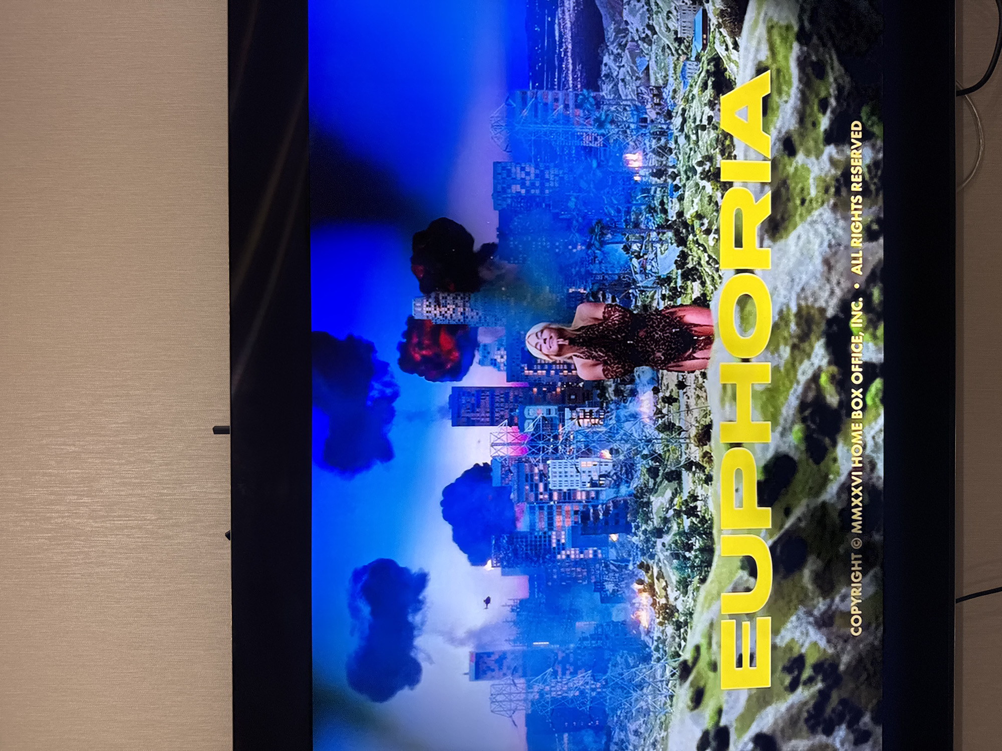 The image shows a television screen displaying the title card for the TV series "Euphoria." The background features a city in apparent ruins, with smoke and destruction visible. A blonde woman is visible in the center, posing with a smile. The word "Euphoria" is superimposed in large yellow letters on the right side. Copyright information is printed below the title. The city is otherwise unidentifiable.