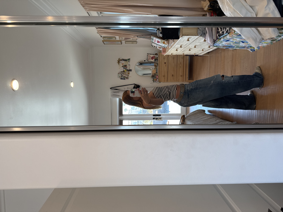 The image displays a young female, identified by her reflection in a large mirror, taking a selfie within a residential interior in Los Angeles, United States of America. The subject, possessing light brown hair, is depicted in a left profile stance, holding a light-colored smartphone in her extended right hand towards the mirror. She is dressed in a black and white striped strapless top, distressed blue jeans with a torn knee, a studded belt, and light-colored slip-on footwear.
The reflected room exhibits white walls, crown molding, wooden flooring, and a white ceiling with two recessed light fixtures. Furniture visible includes two light-toned wooden dressers; the left dresser is topped with a circular mirror and a collection of papers and photographs affixed to the wall above it, while the right dresser holds framed pictures, a black object, and other small items. Framed artwork is displayed on the wall to the right of the dressers, alongside drawn-back pink curtains. A bed with a striped mattress or cover is partially visible on the left side of the reflection. Through a set of glass double doors in the background, bright sunlight and outdoor vegetation, including palm trees, are discernible.