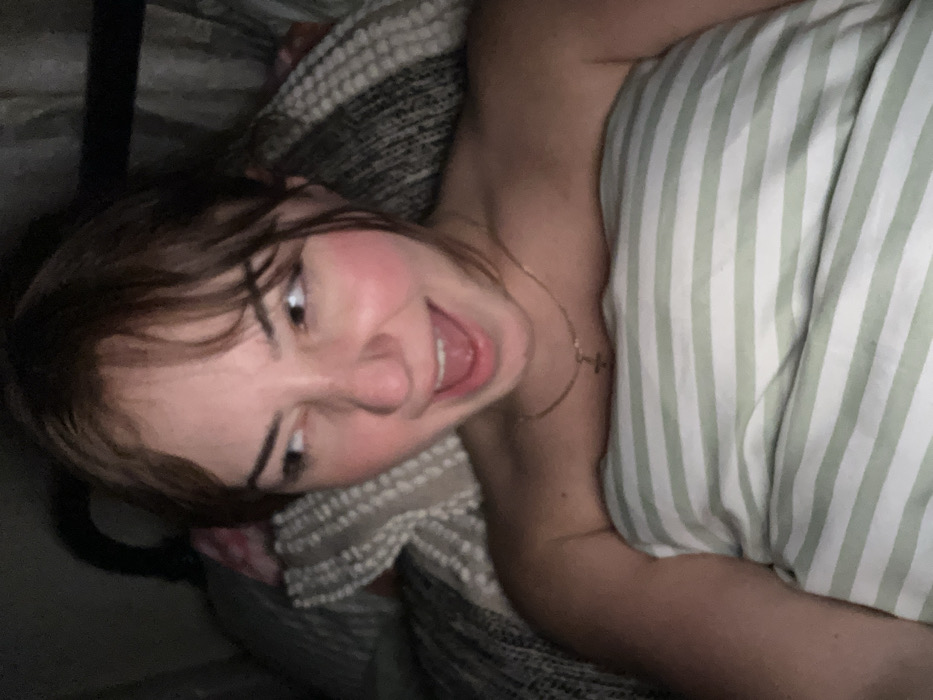 A fair-skinned young woman with brown hair and bangs smiles widely, exposing her teeth. She is lying in bed, with a striped pillow or blanket beside her. She wears a delicate necklace with a cross pendant. One arm is visible, extending towards a striped bedsheet. The background appears dark, possibly due to low lighting in the room.